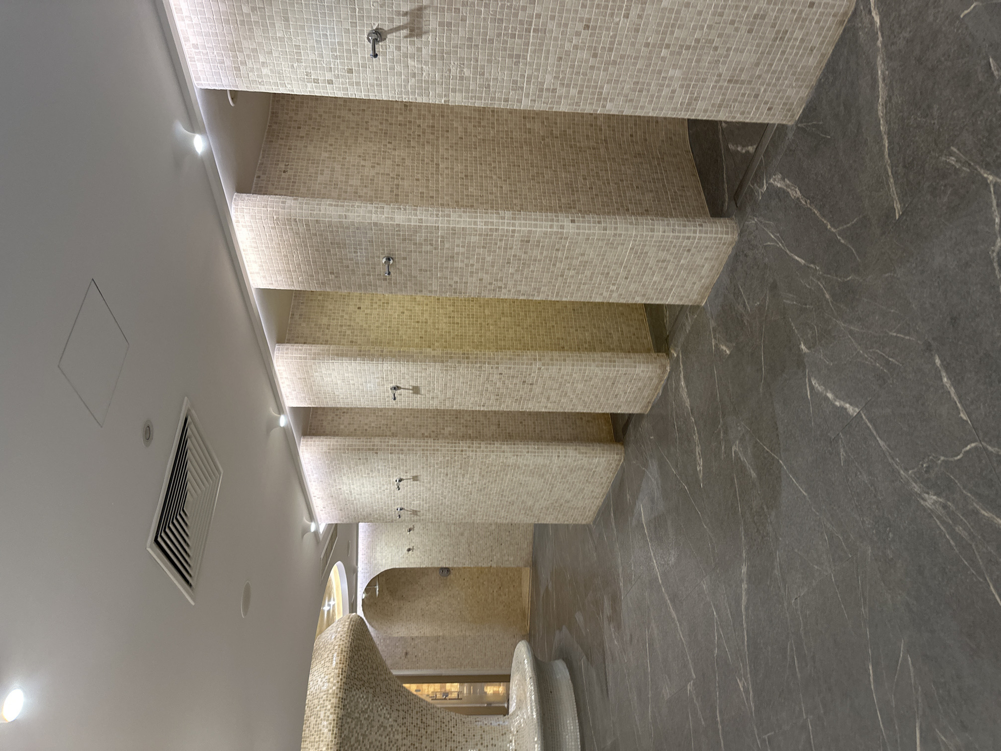 The image shows an interior view of a public bathing area, possibly a thermal bath or spa. The area features multiple shower stalls lined with small, square, beige-colored mosaic tiles covering the ceilings and partial walls of each stall. The floor is covered in large, dark gray tiles with lighter gray veining, resembling marble. The left side of the image shows a white wall with recessed lighting and an air vent. There are also rounded structures covered in mosaic tiles, possibly part of a seating or decorative element. The scene appears to be well-lit and clean. Location in Kazanlak, Bulgaria is not discernible in the image.
