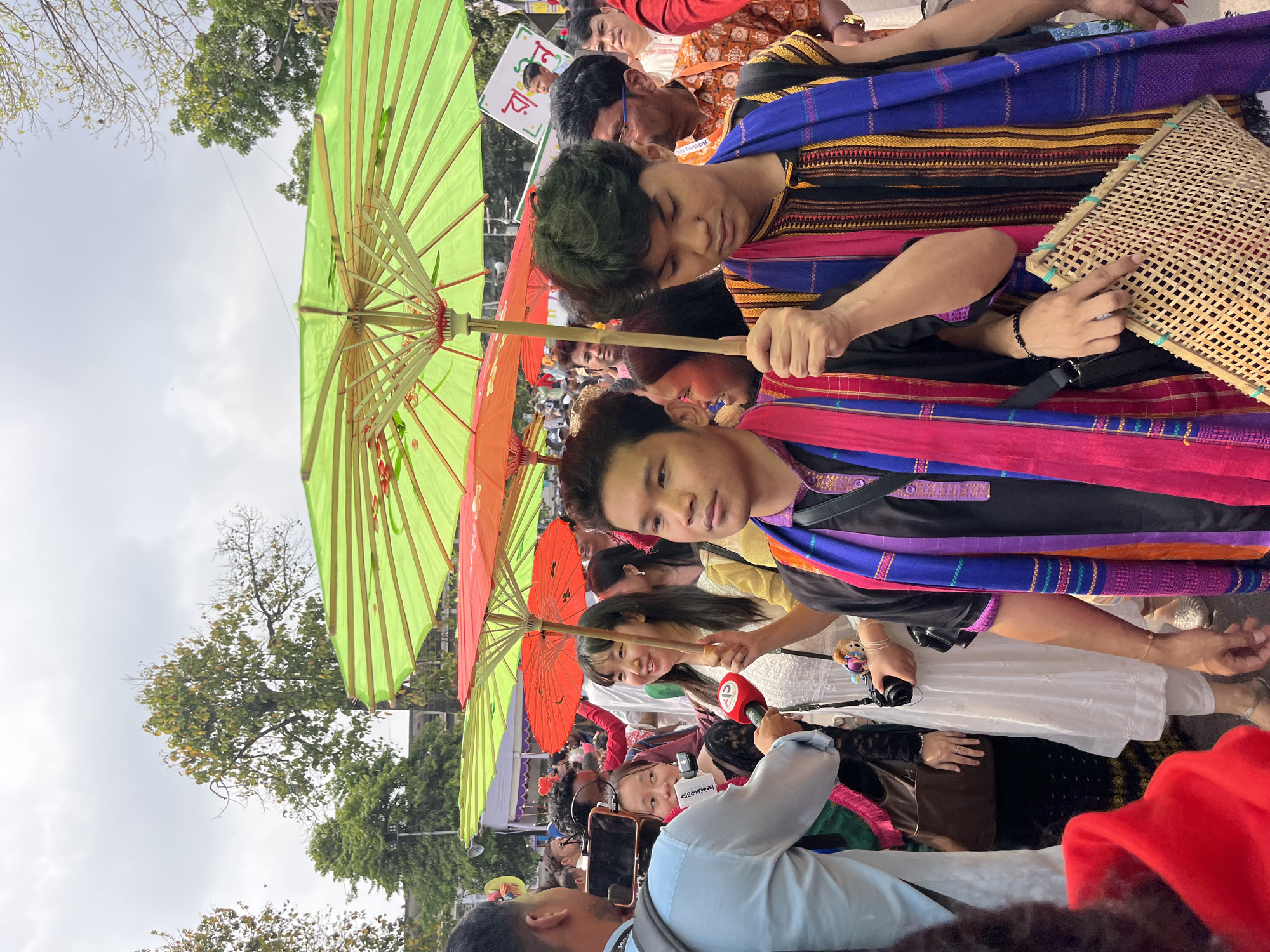 A street-level scene in Dhaka, Bangladesh, shows a crowded street celebration. Multiple individuals are visible, with some holding vibrant, colorful umbrellas, primarily green and orange. People are dressed in festive attire, including patterned shawls and traditional clothing. One person holds a woven, rectangular object, possibly a fan or mat. Some individuals appear to be looking at the camera, while others are engaged in conversation or observation. The environment suggests a public gathering, possibly a cultural or religious event. Overhead are trees and a somewhat overcast sky.