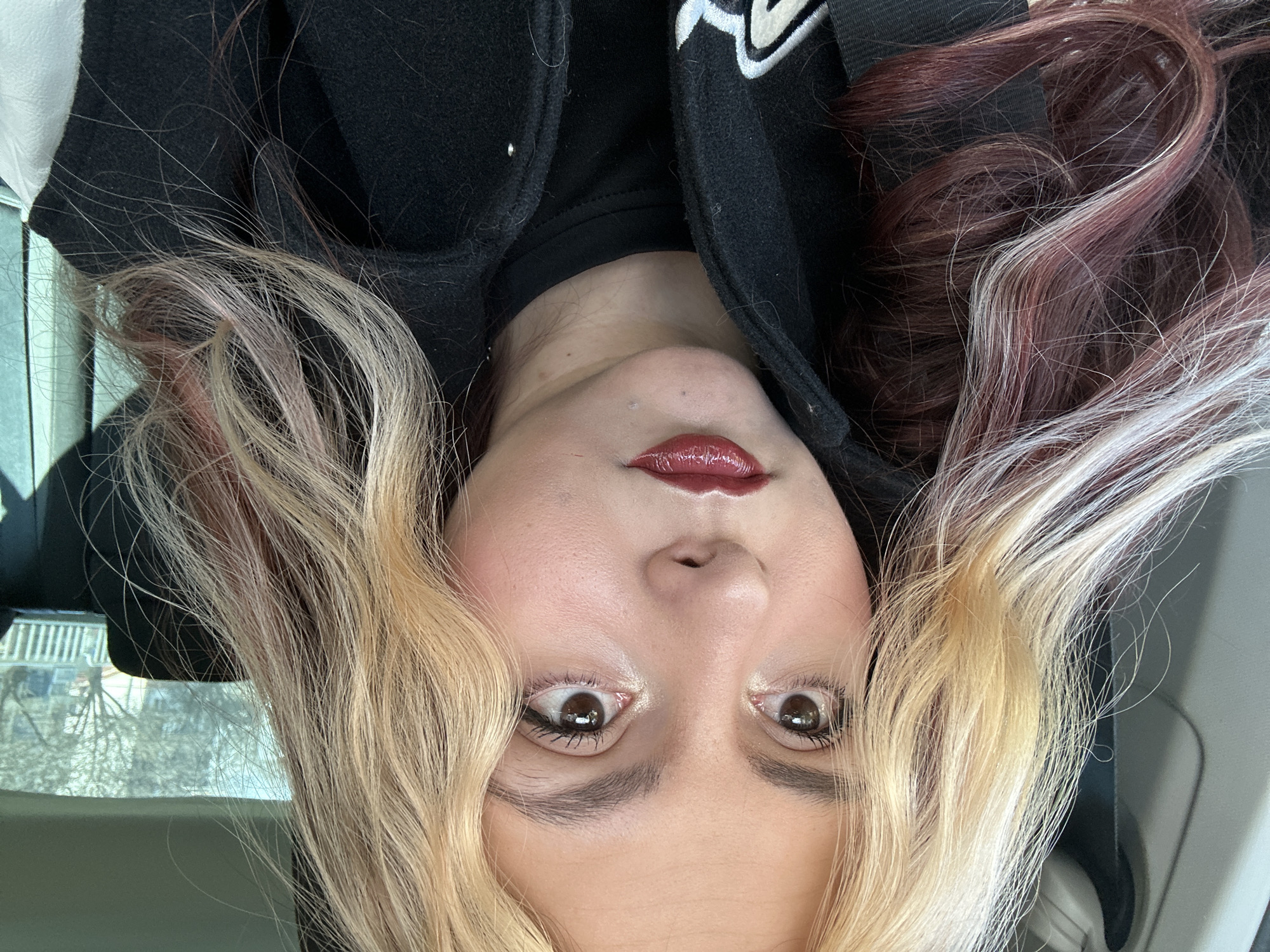 A close-up selfie of a person with dyed blonde and reddish-brown hair. The person has light skin, dark eyes, and dark red lipstick. The person is wearing a black jacket with a white design on the shoulder. The location appears to be inside a vehicle, possibly a car. The image doesn't provide specific details to identify the city as Sofia, Bulgaria, there is no information in this image to identify the city or country the subject is located in. The image is rotated to the right.