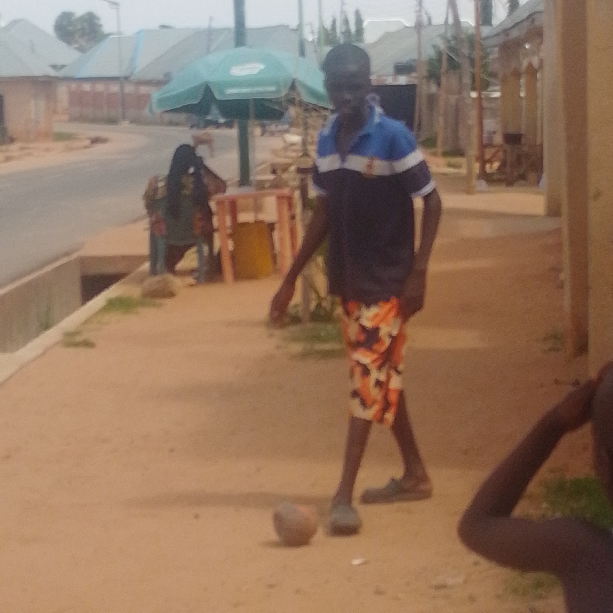 This is a medium-shot, slightly blurry photograph of an outdoor scene. The main subject is a young person wearing a blue and white striped shirt and patterned shorts. They are standing on a dirt path, looking down at a ball at their feet, and appear to be in the process of playing with it. To the left, in the background, a person is seated behind a makeshift stall with a green umbrella. Various structures, possibly buildings with corrugated metal roofs, are visible further in the background. The ground is sandy and uneven, with some sparse vegetation. The lighting suggests it is daytime, possibly overcast or late afternoon, as there are no harsh shadows. The overall atmosphere appears to be casual and everyday, likely in a rural or semi-urban setting. The image quality is low, with visible noise and lack of sharp detail, which obscures specific location cues and fine details of the subjects and their clothing. No text is discernable in the image.