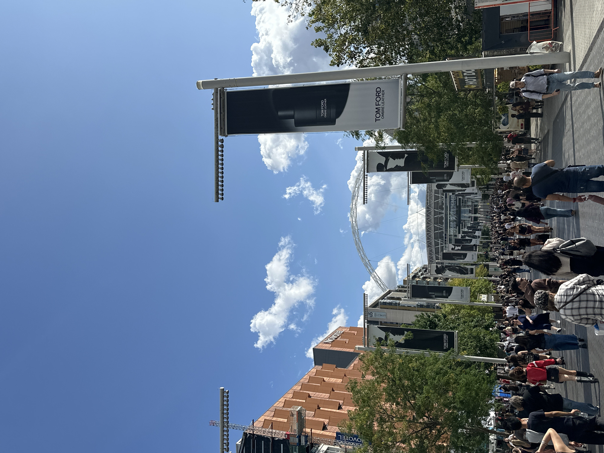 The image depicts Wembley Stadium, a prominent landmark in London, United Kingdom. A large crowd of people is walking down a wide pedestrian walkway. Overhead, banners featuring "Tom Ford Ombre Leather" are displayed on lampposts, advertising the fragrance. To the left of the walkway, a building with distinctive copper-colored facade, identified by a "NOVOTEL" sign, is visible. Further in the distance, the iconic arch of Wembley Stadium can be seen. The sky is blue with scattered white clouds, indicating fair weather. Several food stalls are present on the right side of the walkway, one with a "HOT DOG" sign. The scene suggests a busy event day, possibly before or after a concert or sporting match at the stadium.