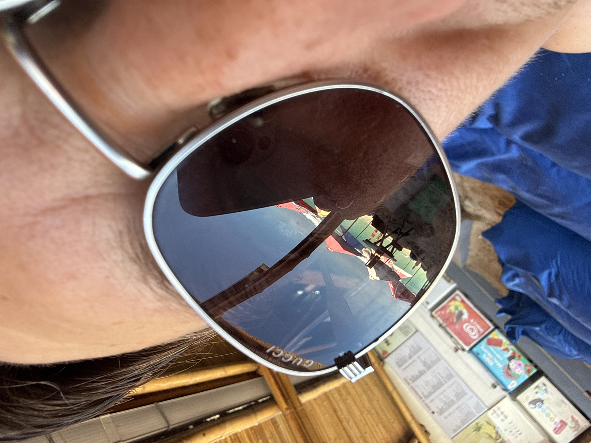 A close-up shot shows a person wearing sunglasses with the brand name "GUCCI" visible. The reflection in the sunglasses depicts a beach scene in Kuta Selatan, Indonesia. Visible elements in the reflection include beach umbrellas, tables, chairs, and the beach itself. The person's skin is visible around the glasses. In the lower right of the image is a blue material, a bamboo structure, a printed document, and ice cream treats.