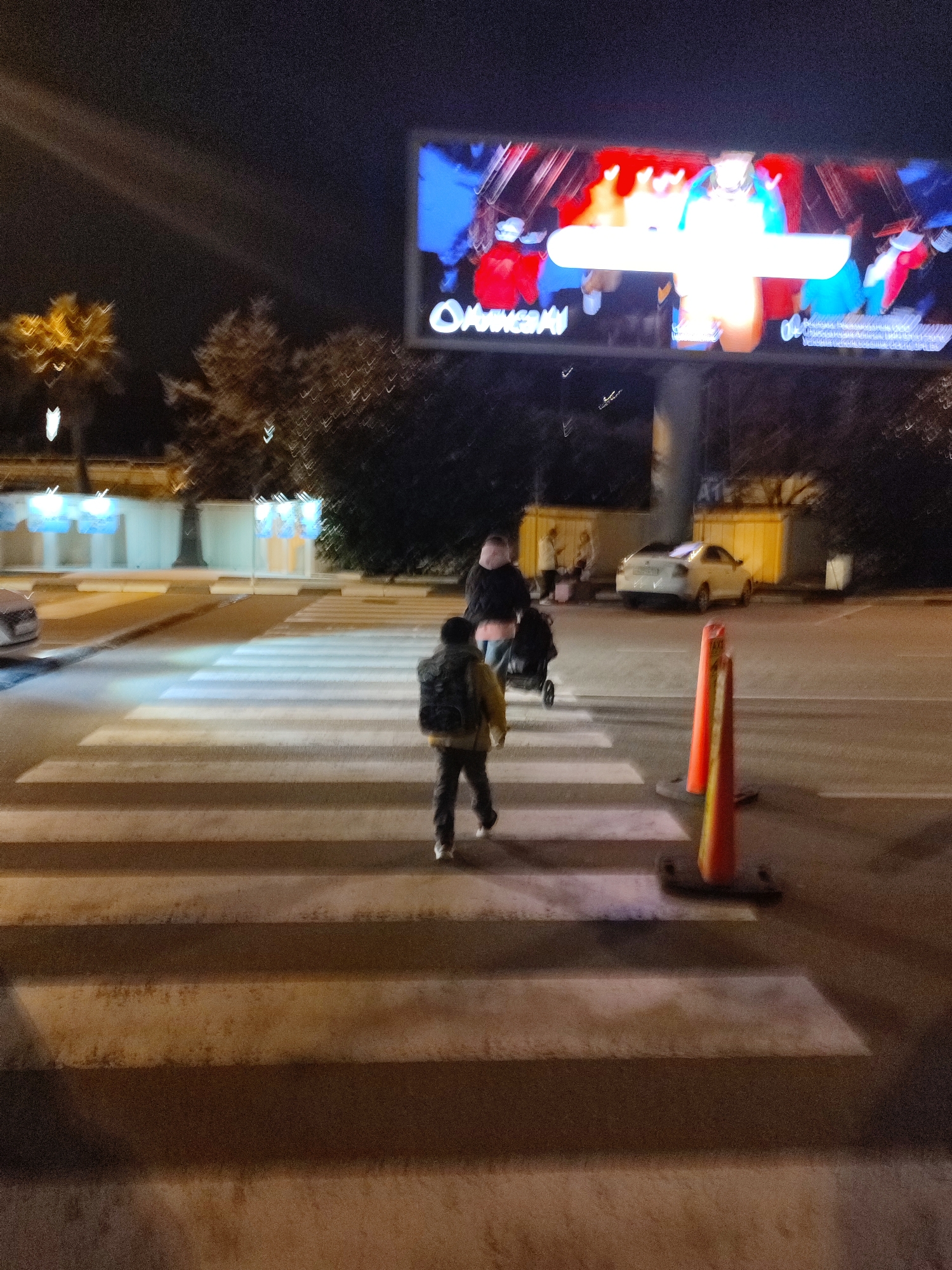 The image depicts a nighttime urban street scene at a pedestrian crossing in Sochi, Russia. In the midground, a child or young person wearing a dark backpack and light top walks from right to left across the striped pedestrian crossing. To their right and slightly behind, an adult in a dark jacket pushes a dark baby stroller, moving from left to right on the crossing. Two orange traffic cones with black bases stand on the asphalt road to the right of the crossing.
In the background, a large, illuminated digital billboard on a tall pole displays abstract, vibrant content featuring red, blue, and white colors, with the partially visible Russian text "ОЛИМПИАДА" (Olimpiada/Olympics) visible on its lower section. A white sedan is parked on the right side of the street, near a yellow-toned building or wall, with two additional indistinct figures visible on the sidewalk beyond it. On the far left, a light-colored vehicle is partially visible. The environment includes dark trees, some resembling palm trees on the left, and various illuminated structures and facades lining the street. Artificial street lighting illuminates the wet-appearing asphalt road and the pedestrian activity. The overall image exhibits motion blur and characteristics of low-light photography.