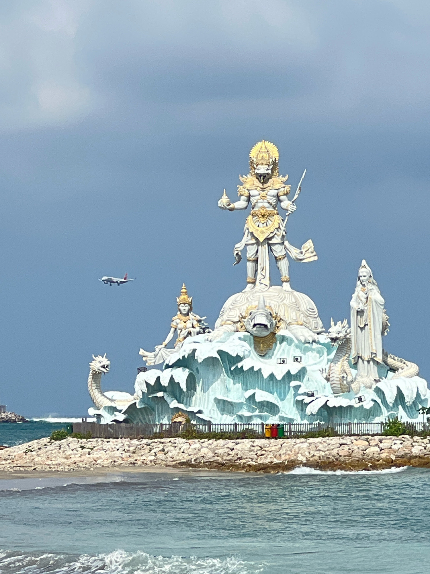 An image captured from the sea shows a large, elaborate statue on a rocky outcrop, likely in Kuta, Indonesia. The statue is a multi-tiered structure depicting mythological figures. At the top, a prominent figure, possibly Vishnu in his avatar as Narasimha, stands on a giant turtle, holding a spear and a cup. Flanking this central figure are smaller statues of a seated woman with a headdress on the left and a standing robed figure with a serene expression on the right. The entire structure is adorned with decorative elements resembling waves and is guarded by two serpentine dragon-like creatures on either side. In the background, an airplane is visible in the sky, and the sea with gentle waves occupies the foreground. The sky is overcast with dark clouds.
