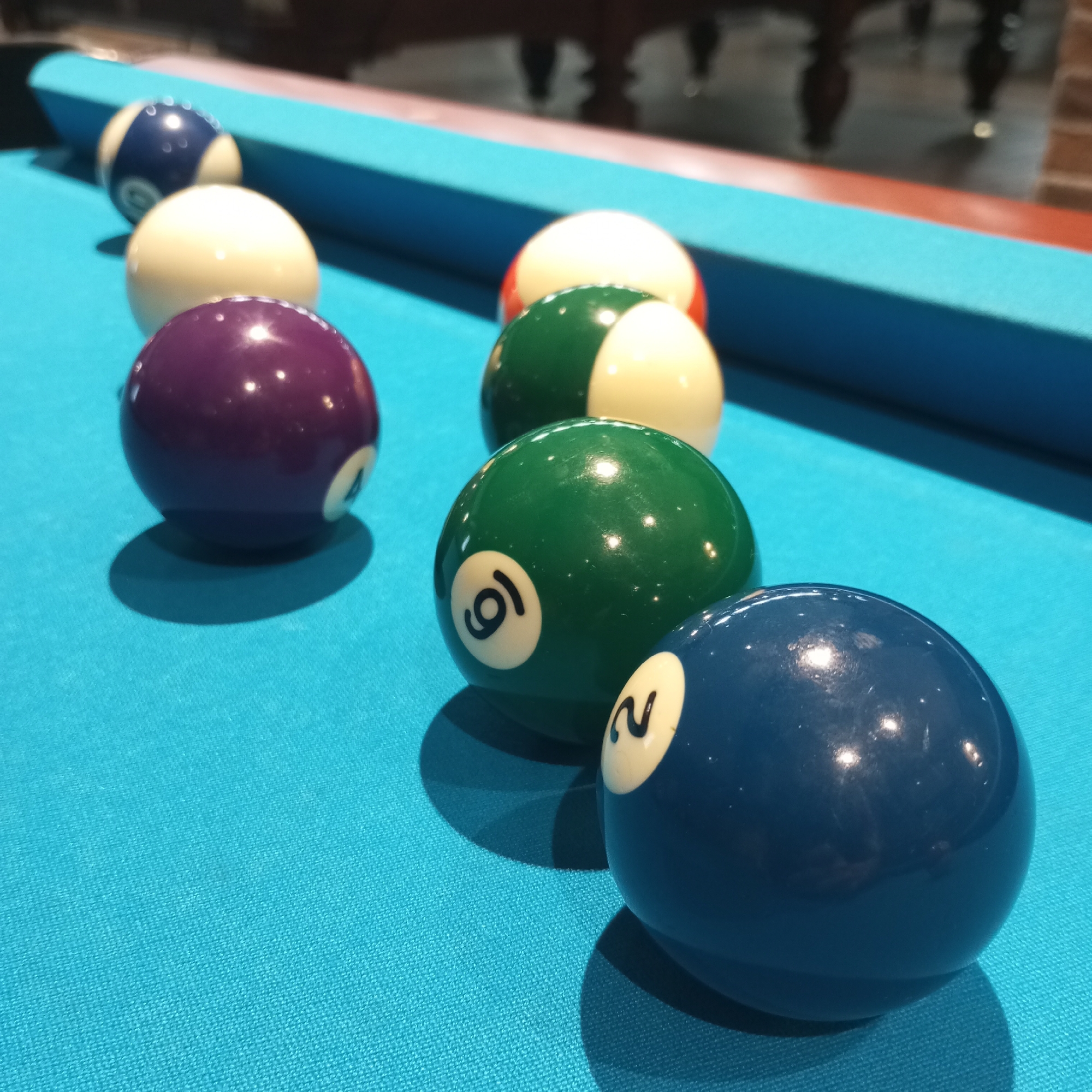 A close-up, eye-level shot shows several billiard balls arranged diagonally on a teal pool table. From front to back, left to right, the visible balls are a solid blue ball with a white circle containing the number "2", a green ball with a white circle containing the number "9", a solid purple ball with a white circle containing the number "4", a white ball, a striped blue and white ball with a white circle containing the number "1", and a ball that is mostly white with a red stripe. The balls are well-lit, with reflections visible on their surfaces, and cast distinct shadows on the felt. The background is out of focus, showing the legs of a wooden pool table and a hint of a brick wall. The setting appears to be an indoor recreational facility. The image does not provide any information about the city of Izhevsk, Russia.