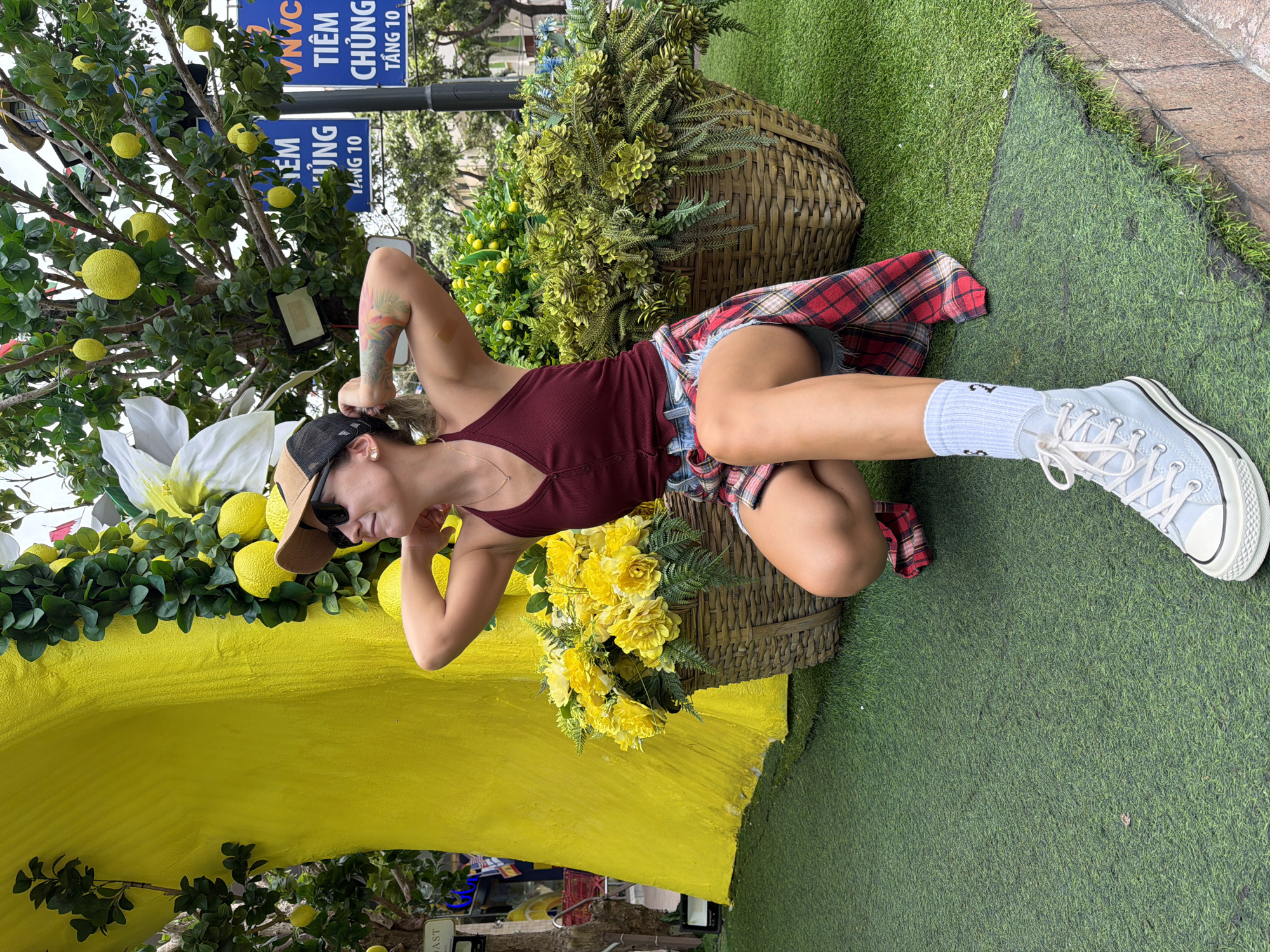A young woman with blonde hair pulled back, wearing a maroon tank top, denim shorts, a plaid shirt tied around her waist, a tan baseball cap, and light blue high-top sneakers, crouches on artificial turf. She is positioned in front of a large, bright yellow, tree-like sculpture adorned with artificial lemons and greenery. Two large wicker baskets filled with yellow flowers and lush green plants are situated behind her, one to her left and one to her right. In the background, signs in Vietnamese and English are visible, with one sign reading "TIEM CHUNG" (Vaccination) and "TANG 10" (Floor 10), indicating a location in Nha Trang, Vietnam. The scene appears to be an outdoor urban setting, possibly a decorative display in a public area or commercial establishment.