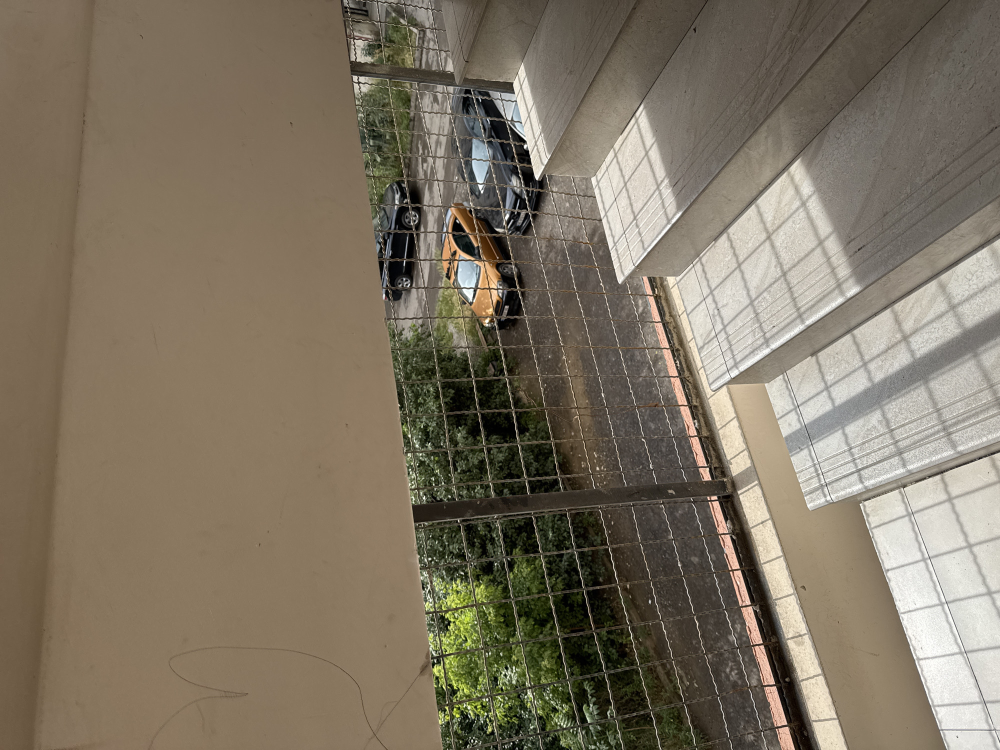 The image shows a view from a concrete staircase overlooking a courtyard with parked cars and trees. The staircase is made of light-colored tiles with visible grid patterns from sunlight and shadows. The courtyard has a paved ground and is enclosed by a metal fence. Three cars are visible: a black SUV in the background, an orange hatchback in the middle, and a black sedan in the foreground. Dense green trees form a backdrop behind the fence. The scene appears to be outdoors, possibly in a residential area.