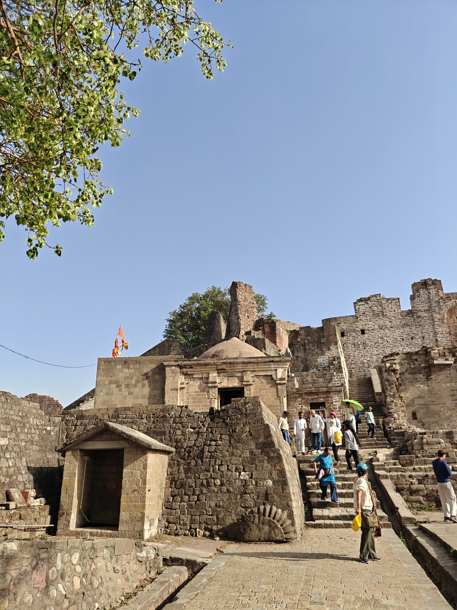 The image shows a section of the Rohtas Fort, a historical fort located near the city of Kaswara, in the Jhelum district of Punjab, India. The architecture is primarily stone-based, and the overall fort is well maintained with some portions showing signs of aging. A group of tourists is seen walking up the stone staircase. The scene is set in daylight with a clear blue sky. A tree is visible in the upper left corner of the frame. A small room-like structure stands near the base of the stairs. A flag is seen on the highest platform of the fort. The setting seems to be outdoor during the day.