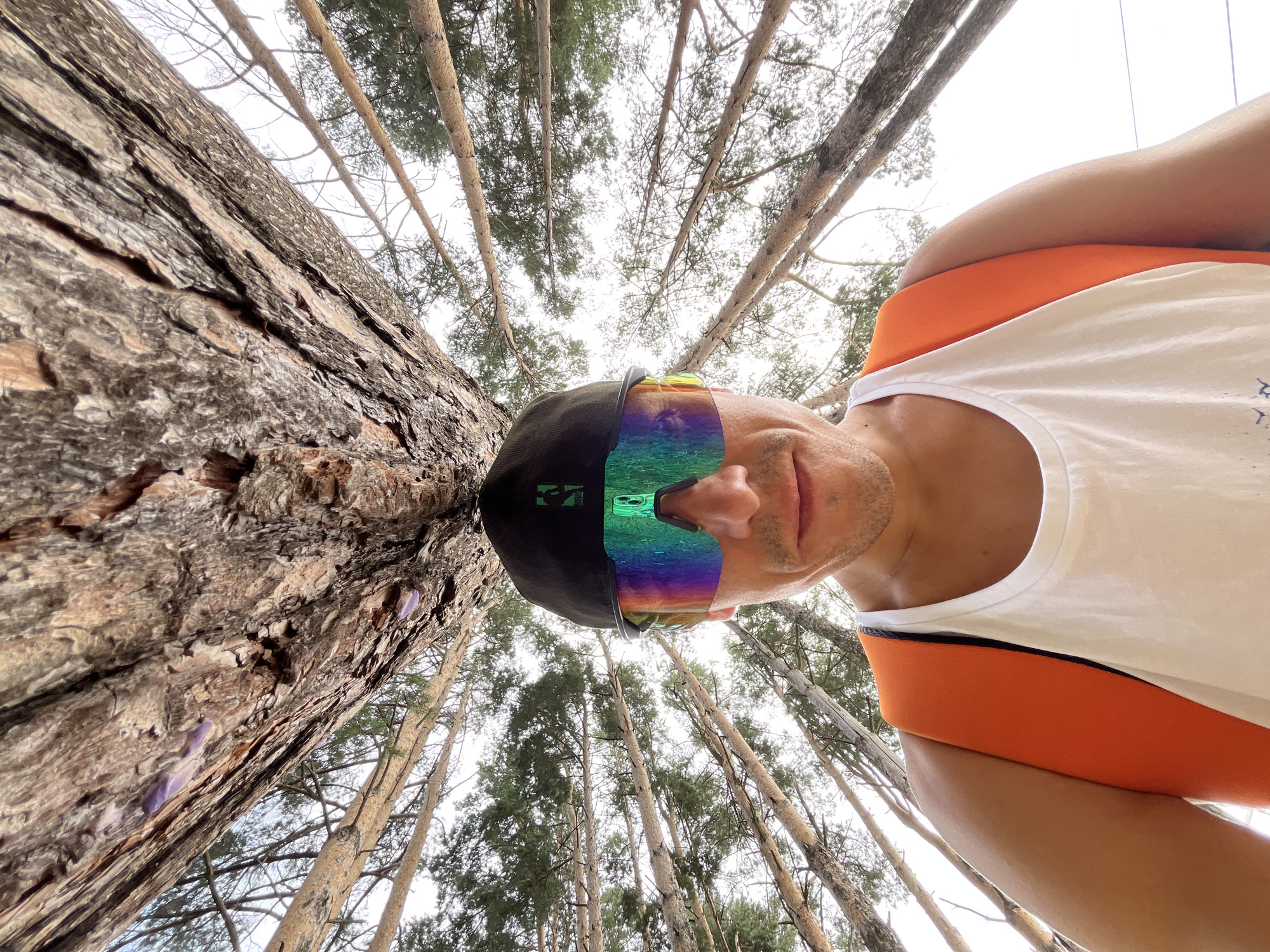 A close-up, low-angle, wide-shot perspective is captured of a person wearing a black cap and mirrored sunglasses, with their face tilted upwards and looking towards the camera. The sunglasses reflect the surrounding forest and sky. The person is wearing a white tank top with orange trim. To the left, the textured bark of a large tree trunk is prominent, filling a significant portion of the frame. The background consists of a forest of tall, slender pine trees, with their branches and needles extending towards a pale sky. The image appears to be taken in a natural, outdoor environment, possibly a forest area within or near the city of Chaikovsky, Russia, given the context provided.