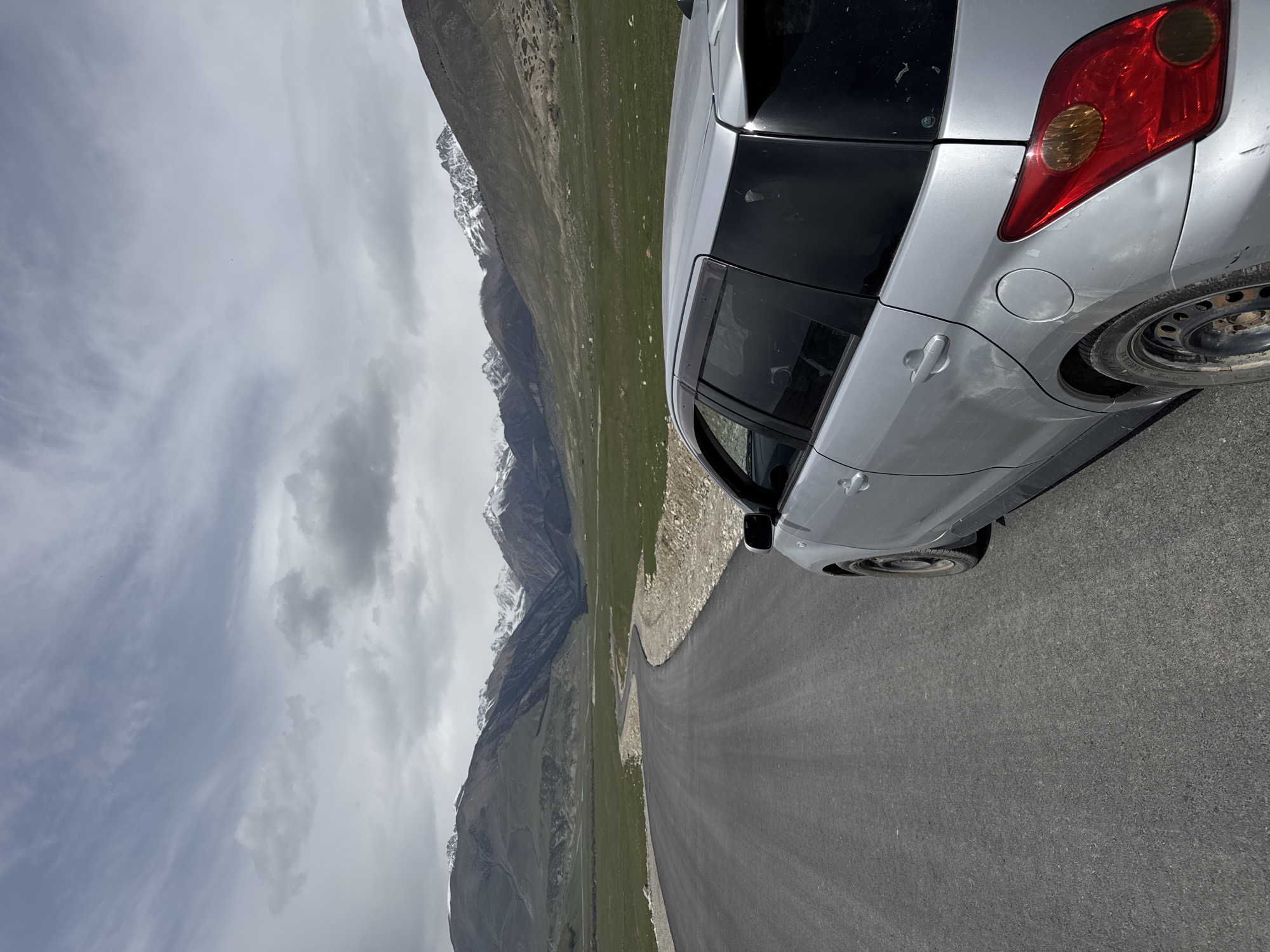 This photograph captures a scenic view of a silver (or light grey) car parked on a paved road, set against a dramatic mountainous backdrop. The car, possibly a compact SUV or hatchback, is positioned on the right side of the frame, showing its rear quarter panel, taillight, and rear wheel. It appears to be a bit dusty, with a possible scuff mark visible near the fuel cap. The road itself is asphalt, curving gently through the landscape.
The scene is dominated by towering mountains in the middle and far distance, their peaks and upper slopes adorned with patches of snow, indicative of high altitude. Below the snowline, the mountains transition into steep, rocky terrain and then into lush green grasslands that fill the valley. The sky is overcast with a mix of light and dark grey clouds, suggesting a cloudy but bright daytime.
The image conveys a sense of a journey or exploration through a rugged and beautiful natural environment, characteristic of mountainous regions. Given the location context of Semenovka, Kyrgyzstan, the landscape is consistent with the country's renowned high-altitude scenery. There are no visible people or discernible text in the image.