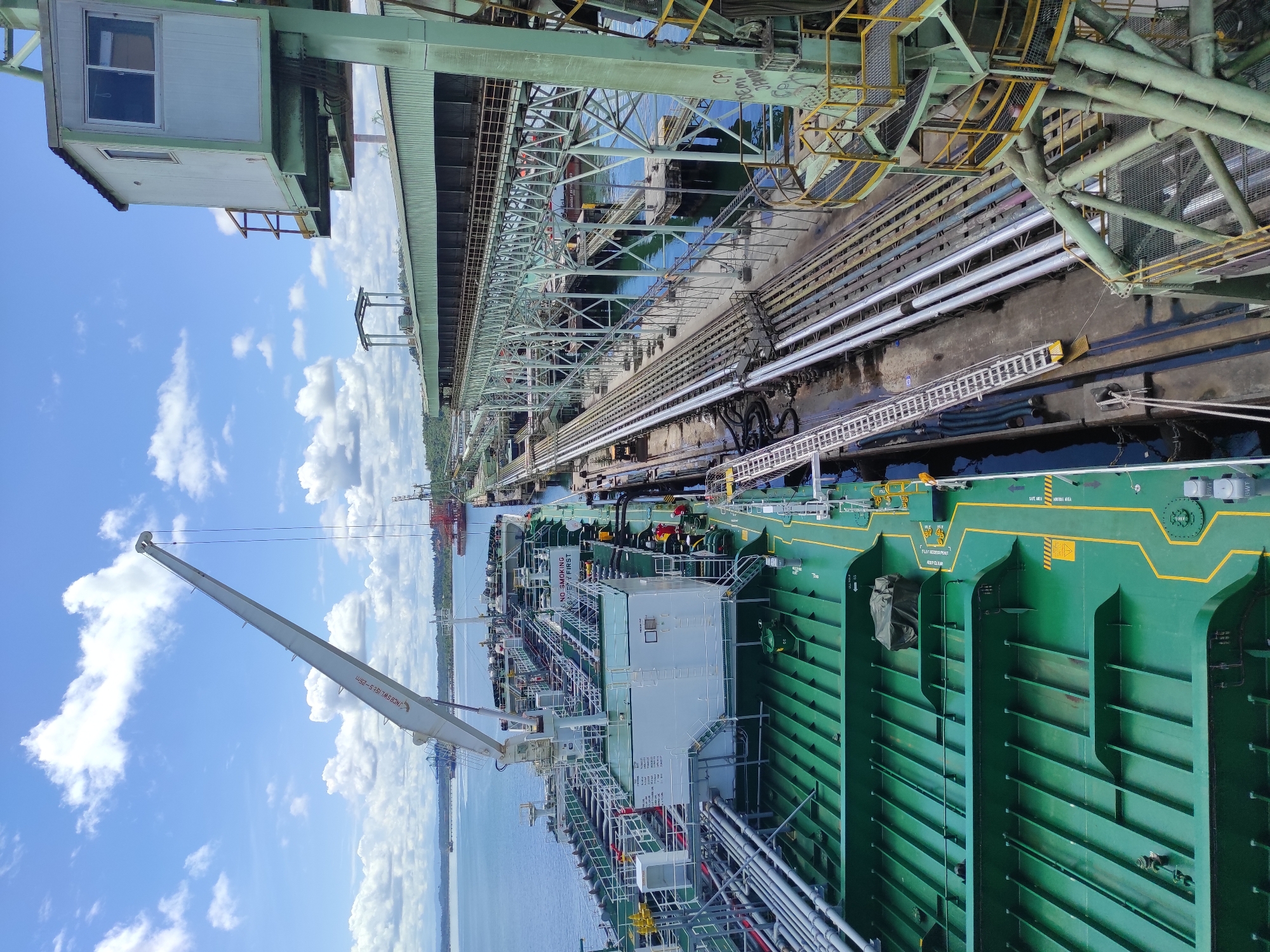 The image shows an aerial view of a port facility with a large cargo ship docked at a pier. The ship, painted green on its deck, is filled with multiple cargo holds. A white crane extends from the ship towards the left side of the frame, with its boom angled upwards. Adjacent to the ship, a complex network of industrial infrastructure is visible, including elevated conveyor belts, pipelines, and support structures. A control booth with windows is perched atop one of the green metal structures on the right. The water in the harbor appears calm and blue. The sky is a vibrant blue with scattered white clouds. The scene suggests an active industrial area engaged in cargo loading or unloading operations.