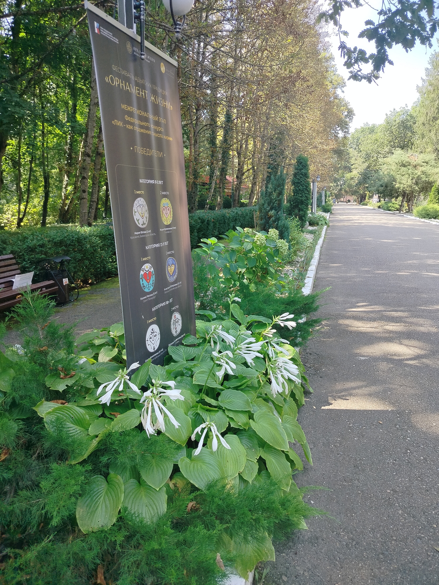 This image depicts a paved pathway in what appears to be a park or public garden. On the left side of the pathway, there is a large, dark banner displaying text in Russian, mounted on a metal pole. The banner details a festival called "Ornament of Traditions" and lists winners and categories. In front of the banner, a variety of plants are visible, including lush green hosta plants with white flowers and feathery green foliage.
To the left of the pathway, further back, a park bench and some decorative metalwork are situated amongst dense green bushes and trees. The pathway itself stretches into the distance, lined with trees on both sides. The overall environment suggests an outdoor public space, likely a park, in a location with abundant greenery and mature trees, indicative of a temperate climate.
While the text on the banner is in Russian and references the "Adyghe traditions," there is no visual information in the image that specifically indicates the location as Krasnyi Most, Russia. The scene is tranquil and suggests a pleasant day outdoors. There are no people visible in the image.