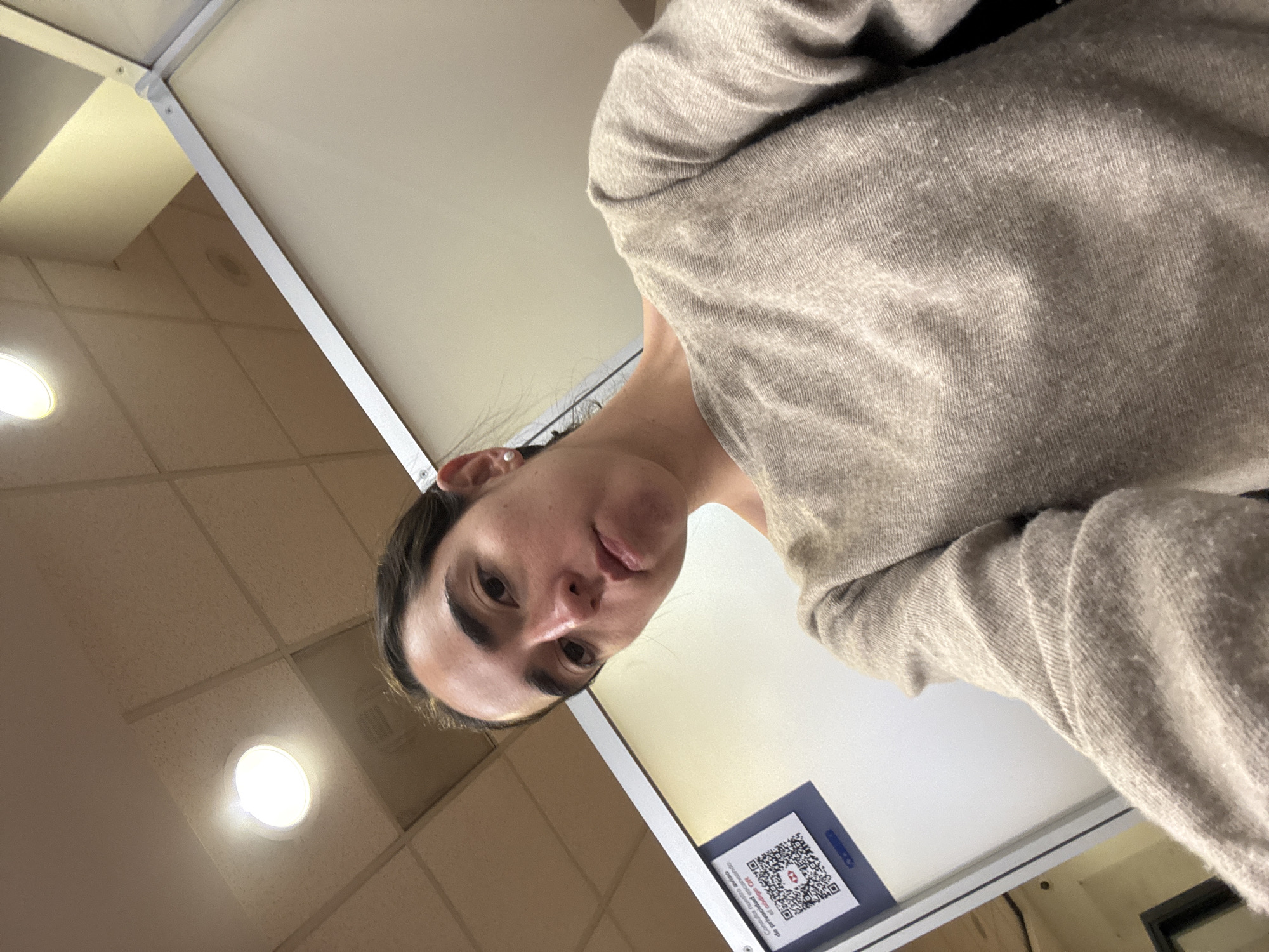 A selfie of a light-skinned woman with dark hair wearing a beige sweater, taken from a low angle. The background includes a drop ceiling with recessed lighting and a white wall. A sign with a QR code is affixed to the wall. Given the HSBC bank logo visible on the sign, and the sign's text in Spanish, the location is likely Mexico City, Mexico.