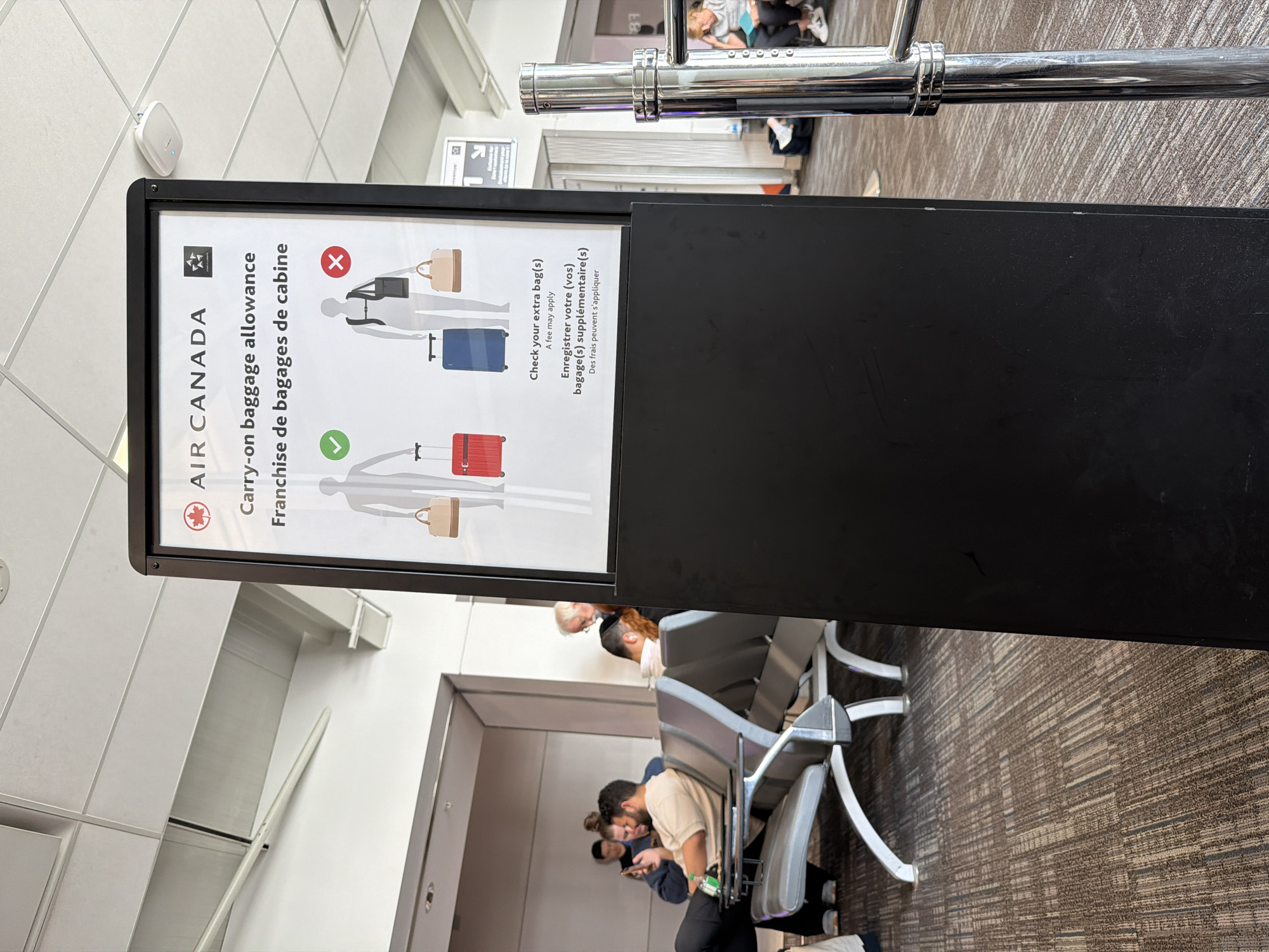 The image depicts a sign for Air Canada's carry-on baggage allowance. The sign is positioned on a black stand in what appears to be an airport terminal. The sign features pictograms illustrating permitted and prohibited carry-on items, along with text in English and French. People are seated in chairs nearby. The location appears to be in Mississauga, Canada based on the Air Canada branding, a Canadian airline.