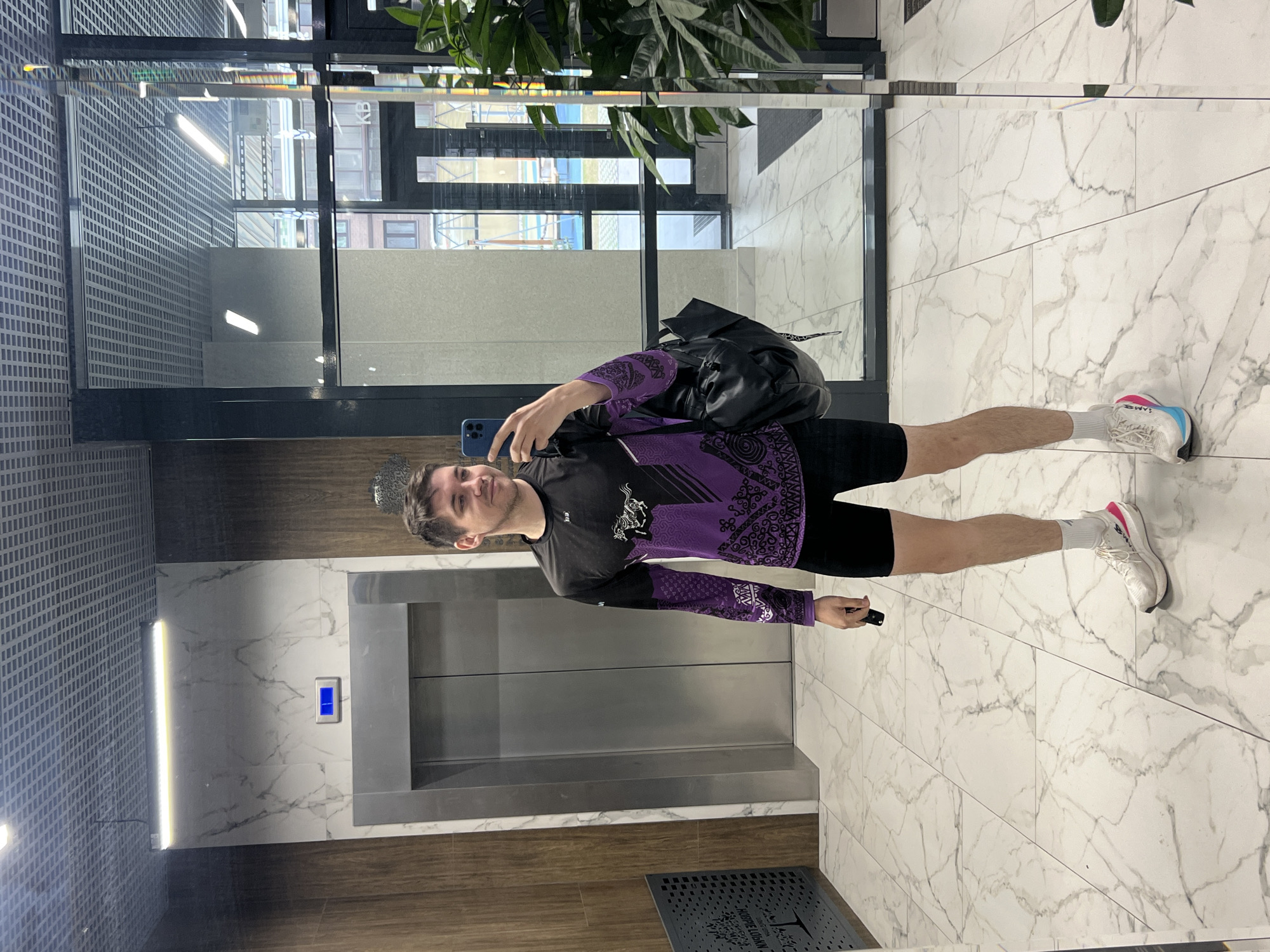 A man is standing in the lobby of a building in Kazan, Russia, taking a selfie with his phone. He is wearing a black and purple long-sleeved shirt, black shorts, white socks, and white running shoes with pink accents. He has a black backpack slung over his shoulder. He is standing in front of an elevator in a modern lobby with white marble floors and walls, and a gray perforated metal ceiling. There is a large mirror on the wall behind him, reflecting the exterior of the building. A plant is visible at the top of the mirror.