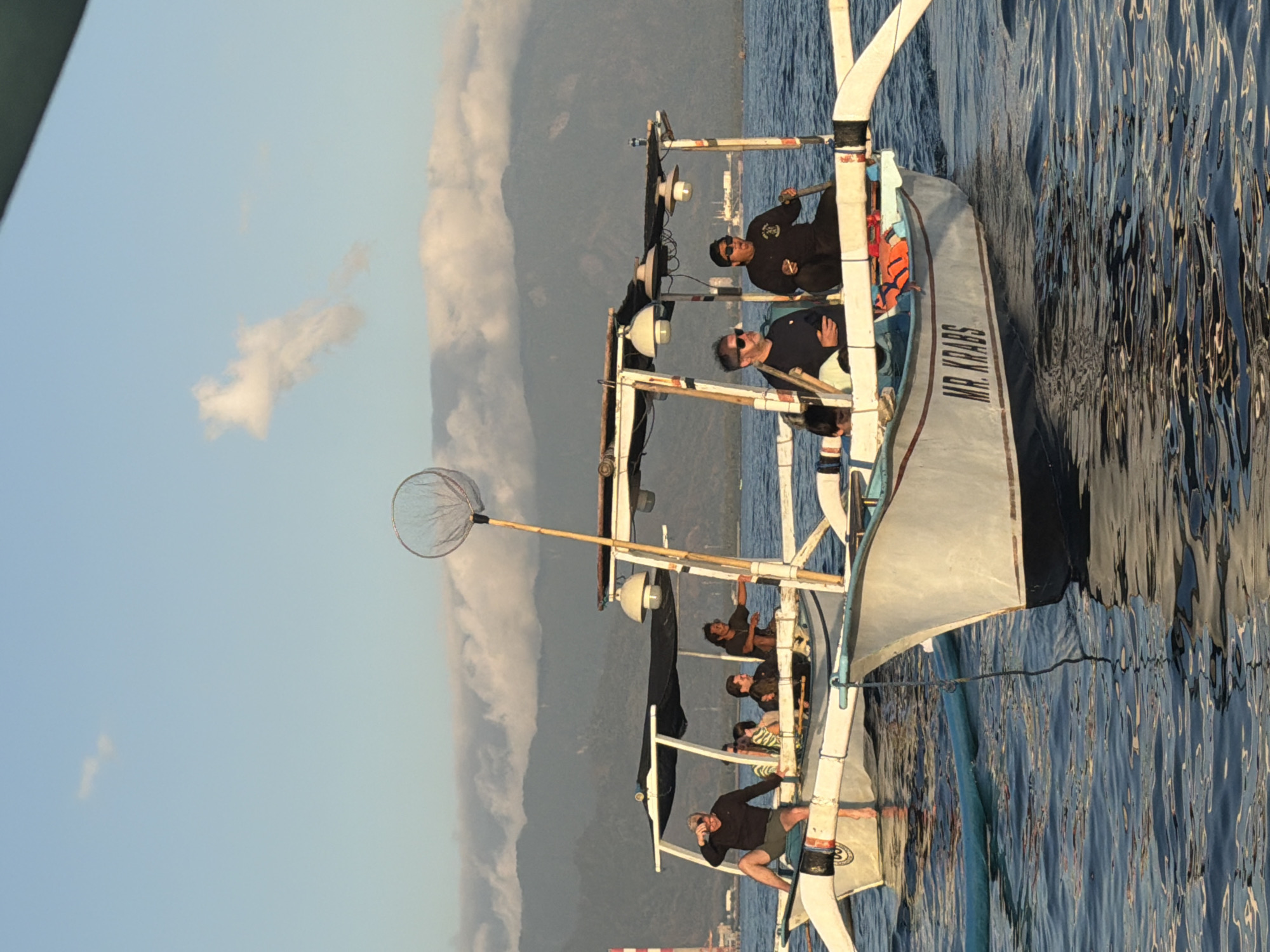 This image depicts two traditional outrigger boats on the water in an unknown location in Indonesia. The boat in the foreground is named "MR. KRABS" and has at least two people visible in it. One person is wearing a black long-sleeved shirt, sunglasses, and a cap, holding an oar. Another person is wearing a dark shirt and also appears to be holding an oar. The boat also has two white lamps mounted on its canopy. The second boat, further in the background, also has a canopy and multiple people on board. One person in this boat is wearing a dark long-sleeved shirt and shorts, with one leg raised as if signaling or gesturing. A net on a pole is extended from this boat. The boats are on calm water, and in the background, there are mountains partially obscured by clouds and a distant industrial structure with a red and white tower. The sky is a clear blue with some wispy clouds.