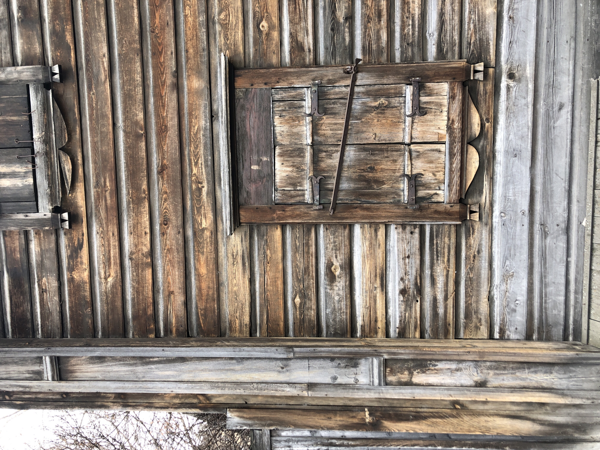 This is an upward-facing view of a weathered wooden structure, possibly a building exterior or interior roof. The location is suspected to be in Krasnoyarsk, Russia.
The scene shows a pattern of vertical wooden planks in various shades of brown and gray. The planks appear to be aged and worn.
There are two wooden framed windows on the wall, one is at the right side of the frame, the second is on the left side. The windows are closed with shuttered wooden panels. The left window is secured with a metal rod.
At the bottom of the frame, there are horizontal planks. The bottoms of several trees can be seen from the bottom of the frame. The weather outside appears to be cold or snowy.