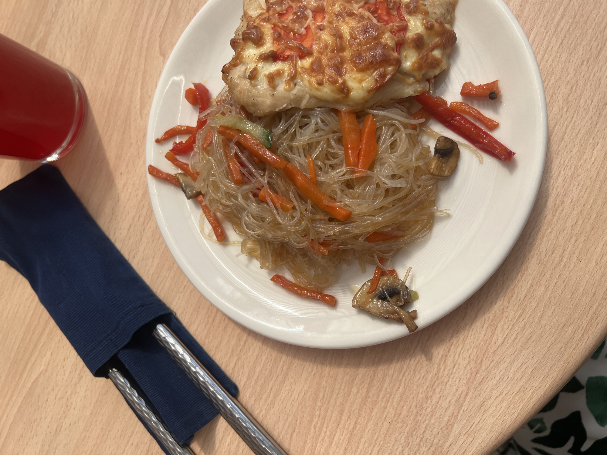 The image shows a plate of food on a wooden table. The plate contains a piece of baked fish topped with cheese and tomato, along with a serving of glass noodles mixed with sliced carrots, red peppers, and mushrooms. Next to the plate is a glass of red liquid, and a pair of chopsticks wrapped in a dark blue napkin. The environment appears to be an indoor setting, possibly a home or restaurant. There are no people visible in the image. The photograph offers no information to connect it to the city of Votkinsk, Russia.
