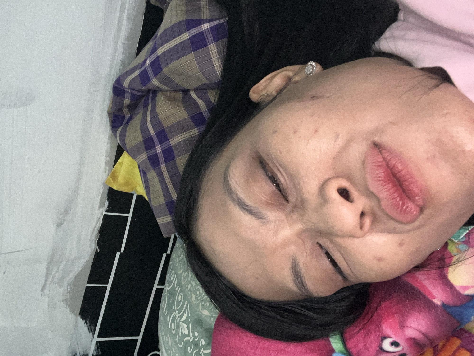 A woman is lying on a bed, her face close to the camera. She has dark hair and is wearing a pink shirt. Her eyes are partially closed, and her lips are pursed. The background shows pillows and a wall with a black and white geometric pattern. The scene is likely in a bedroom in Yogyakarta, Indonesia.
