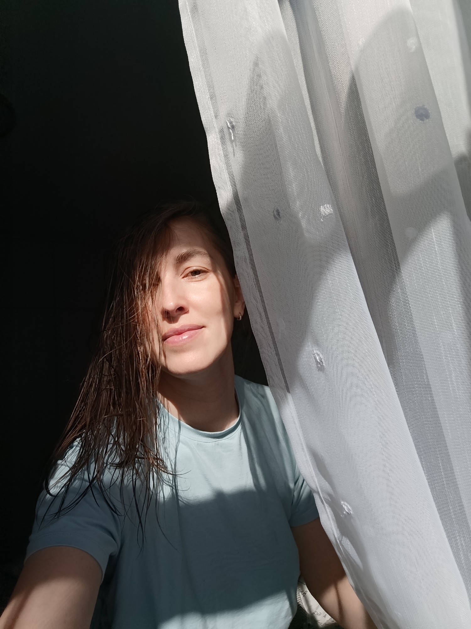 A woman with wet, brown hair is standing in front of a window with white curtains. She is wearing a light blue t-shirt and has a slight smile. Light is coming from the window, illuminating her face and casting shadows on the left side of her face and body. The background is dark. It is not possible to verify the location, Chaikovsky, Russia, from the image provided.