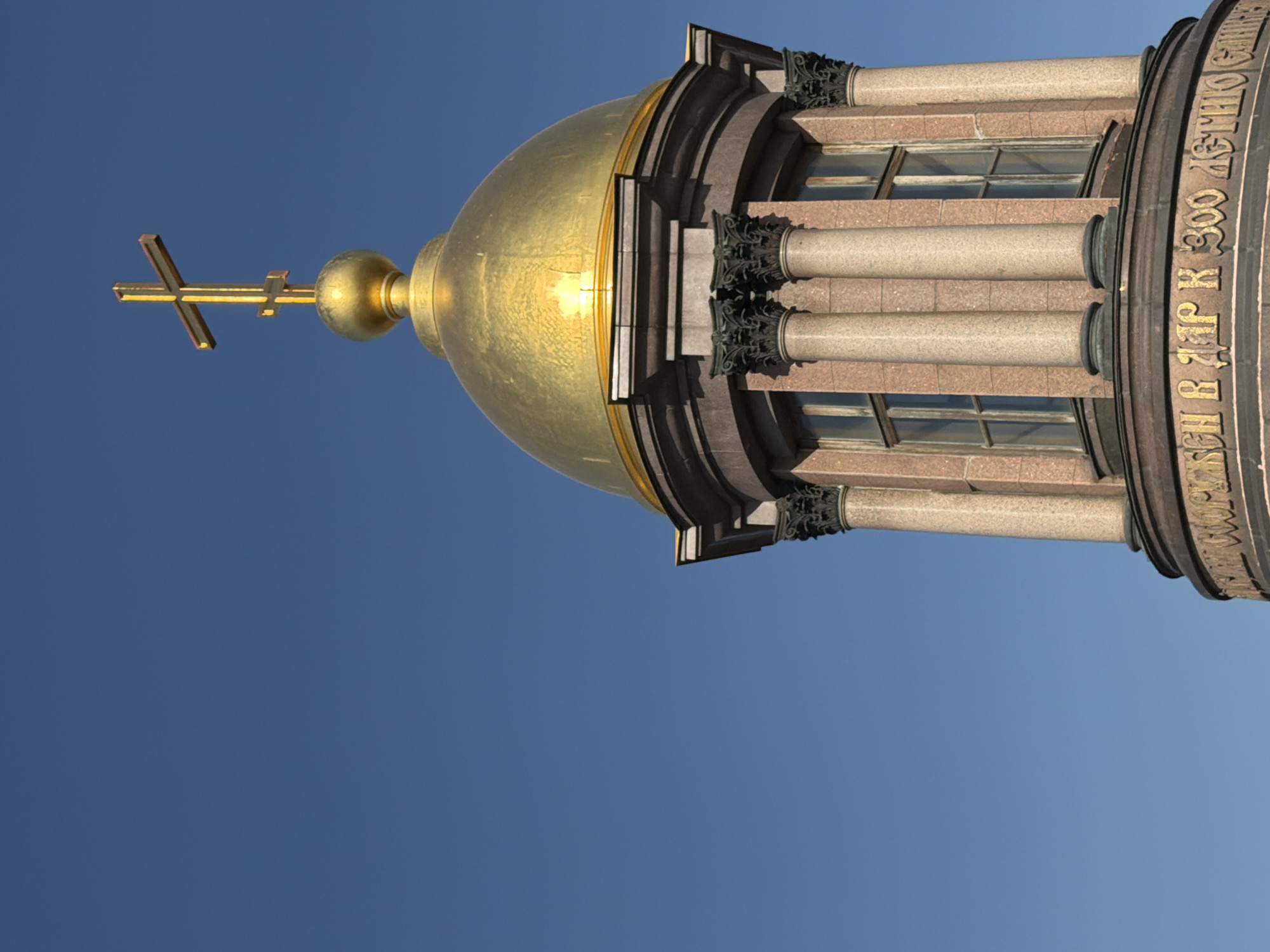The image presents a close-up, upward-angled view of the upper architectural elements of a building against a clear, deep blue sky. The dominant feature is a spherical, golden dome, which exhibits reflective highlights. Topping the dome is a golden Orthodox cross, characterized by a main vertical bar intersected by three horizontal bars: a longer central one, a shorter upper one, and a diagonal lower one.
Below the golden dome is a circular or polygonal lantern structure. This section is constructed from reddish-brown, speckled stone, possibly granite or a similar material. It features multiple slender columns with dark, ornate capitals (likely Corinthian or composite style), positioned between rectangular window openings with dark frames. Around the base of this lantern, a band displays a gilded inscription in Cyrillic script against a dark background, partially legible as "ВЪ ДАР К 300 ЛЕТНЮ" (as a gift to the 300th anniversary).
This architectural feature is identified as the spire's lantern and dome of the Peter and Paul Cathedral, situated within the Peter and Paul Fortress in Saint Petersburg, Russia. No people, other structures, or discernible objects are visible within the frame. The scene is illuminated by natural daylight.