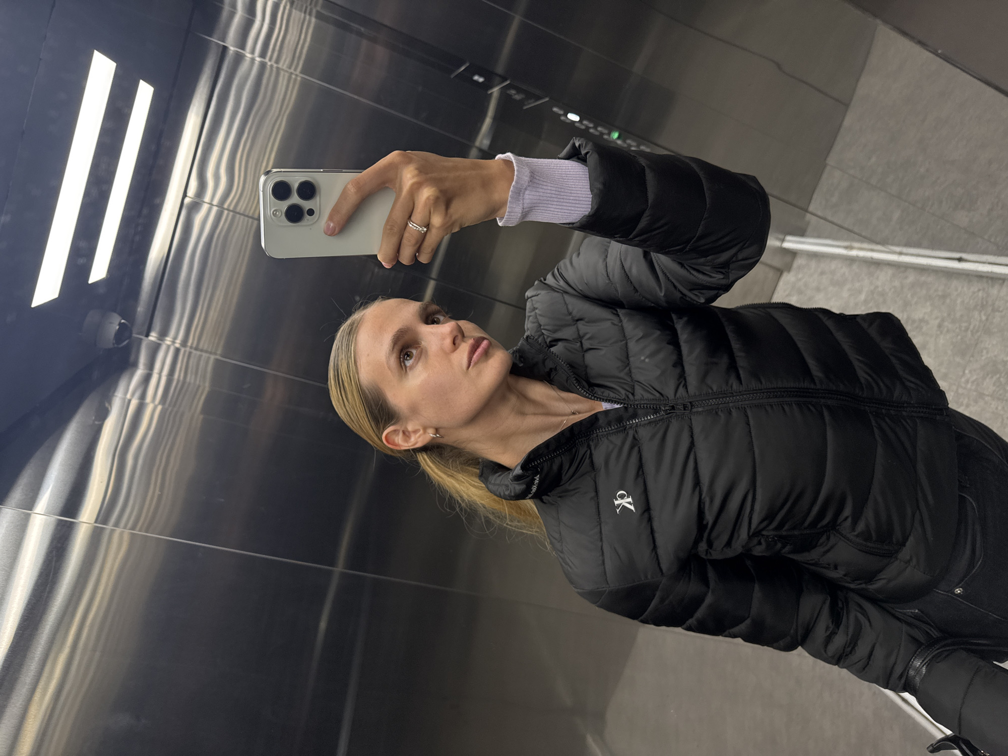 A woman with blonde hair, pulled back, is taking a selfie in a highly reflective indoor environment, likely an elevator. She holds a light-colored iPhone, identifiable by its triple-lens camera system, in her left hand. A ring is visible on her left ring finger, and a light purple ribbed sleeve extends from under her jacket.
She is wearing a black puffer jacket that features a white "CK" logo on the left side of the chest. Her head is tilted slightly upwards, and her gaze is directed towards the phone, indicating she is actively posing for the picture.
The metallic walls of the setting create strong reflections, prominently showing three bright, rectangular light fixtures overhead. A small security camera or sensor is also reflected on the left side of the background. The reflective surfaces contribute to a modern and sleek aesthetic. The overall image has a slightly rotated orientation.