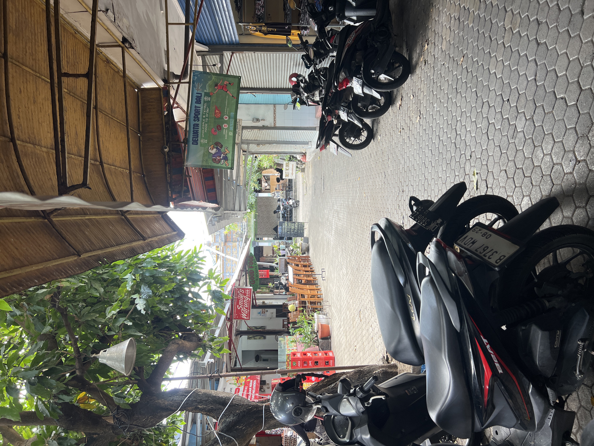 This is a ground-level, eye-level shot of a narrow street or alleyway in Kuta, Bali, Indonesia. The scene is dominated by parked motorbikes on the right side of the frame, lined up along the side of a building. The street is paved with hexagonal cobblestones. On the left, a large tree with lush green foliage overhangs the path, with a hanging light fixture attached to one of its branches. A portion of a motorbike with a black helmet is visible in the foreground on the left.
Further down the alley, several outdoor seating areas with wooden tables and chairs are set up, suggesting a restaurant or cafe. Signs are visible, including one that reads "Smile's Warung" and another with "GOOD FOOD". There are also stacks of red Coca-Cola crates. Buildings line both sides of the alley, with awnings and corrugated roofing visible. A banner with "DEWATA SPORT BALI" is hanging above the shops. The overall impression is of a bustling, informal commercial area, typical of a tourist destination like Kuta. No people are clearly visible in the shot, though some distant figures are suggested in the background. The lighting suggests it is daytime, with sunlight filtering through the trees and awnings.