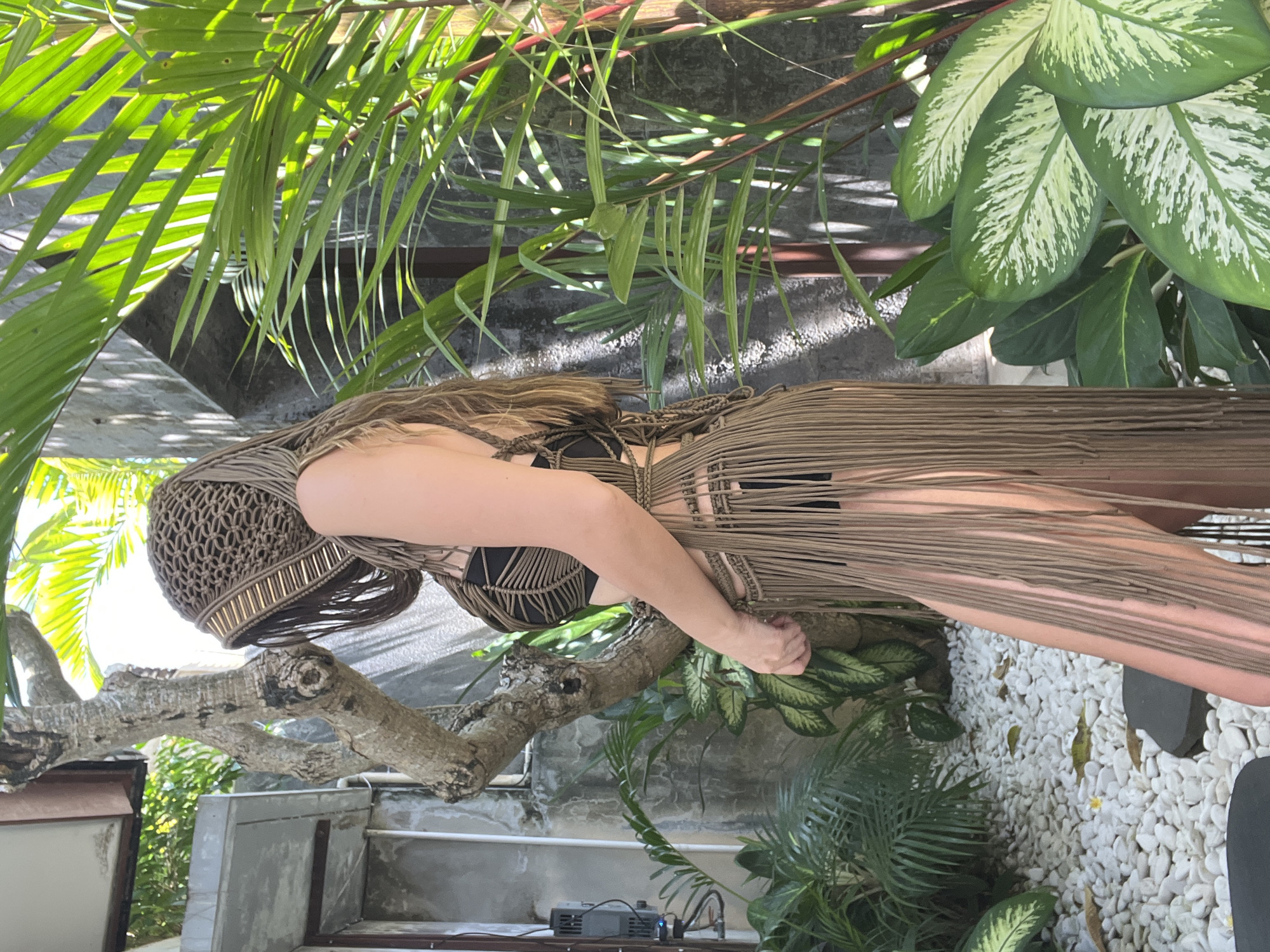 A light-skinned woman wearing a brown macrame dress and a black top poses in an outdoor setting. The location appears to be in North Kuta, Indonesia, based on the tropical foliage and architectural style. She is standing near a weathered tree branch, with lush green plants and palm leaves in the foreground and background. Behind her, there is a wall with a light-colored, textured surface and white pebbles on the ground. In the background, a device with multiple ports is visible on a ledge. The woman's pose suggests she is either leaning against the tree branch or using it for support.