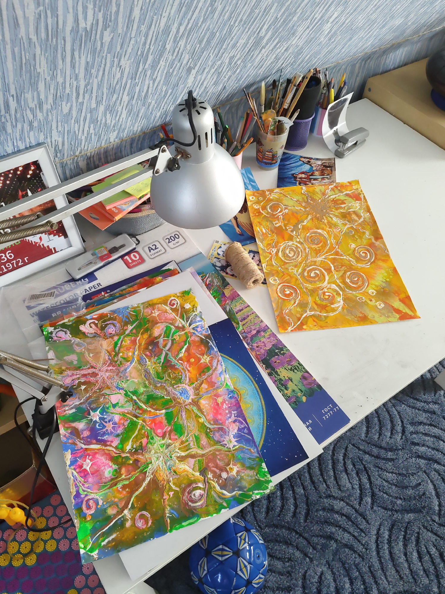 The image shows a desk with art supplies. On the desk, there are several paintings, art brushes in containers, a stapler, a lamp, and reams of paper. The paintings are colorful and abstract, with one being predominantly yellow and orange, and another with a mix of green, pink, and blue. There is also a roll of twine. The desk surface appears to be white and slightly cluttered. The background shows a textured blue wall and a portion of a beige box or shelf. A blue rug with a wave-like pattern covers the floor in the foreground, and a blue and white ball is visible near the bottom of the image. The scene appears to be an indoor art studio or workspace. The image is taken from a high-angle perspective, looking down onto the desk.