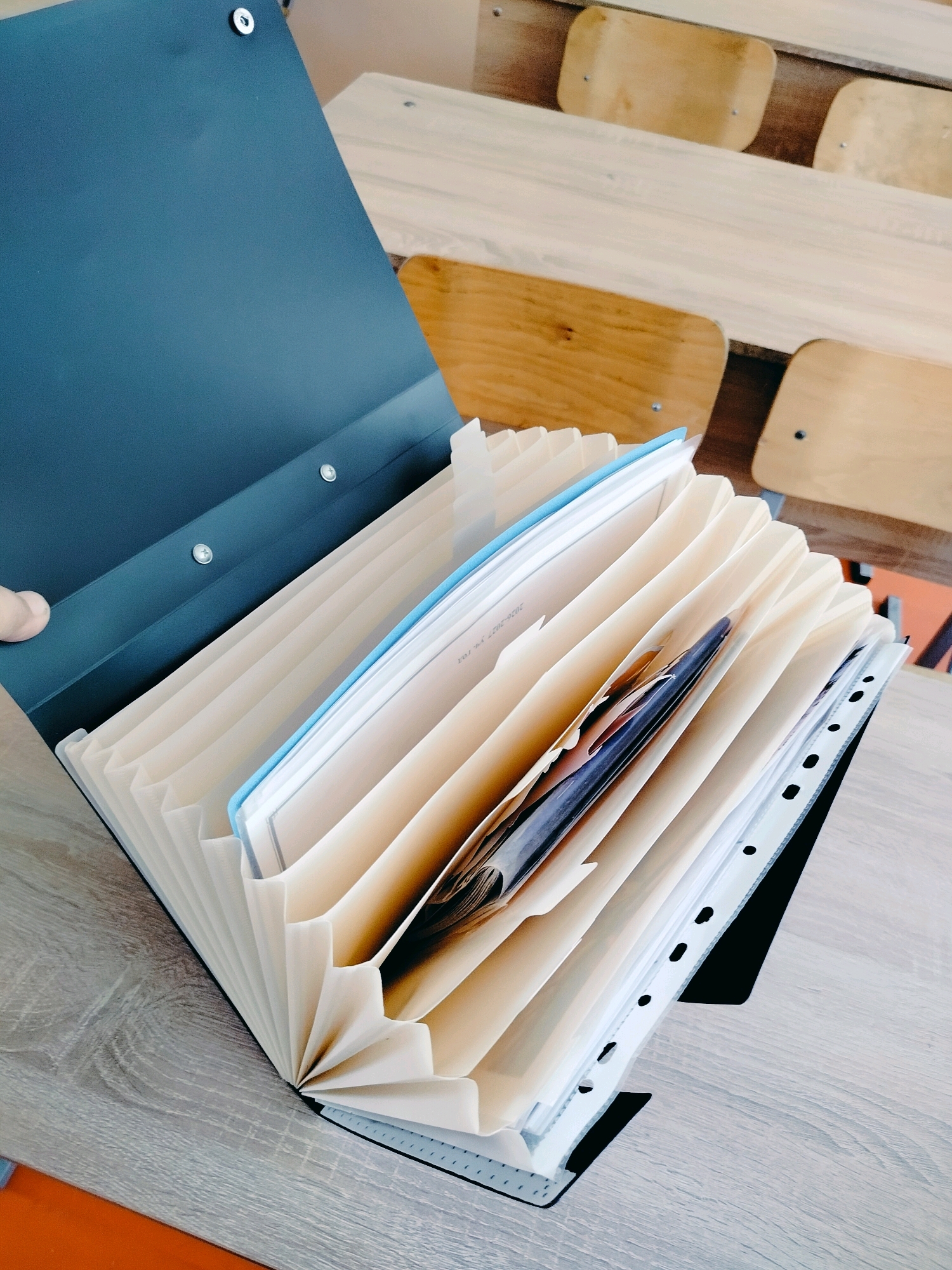 A dark blue accordion file folder lies open on a light-colored wooden surface, possibly a desk or table. The folder is filled with many light-colored paper dividers, some of which contain documents. One divider has a light blue edge. A black binder with punched holes is visible on the right side of the accordion folder. In the background, wooden benches and desks are arranged, suggesting a classroom or educational setting. The lighting is bright, casting subtle shadows. There is no visible person in the image, but a hand is partially visible on the left edge, suggesting someone is interacting with the folder. The location of Көлкент, Kazakhstan is not evident from the visual information in the image.