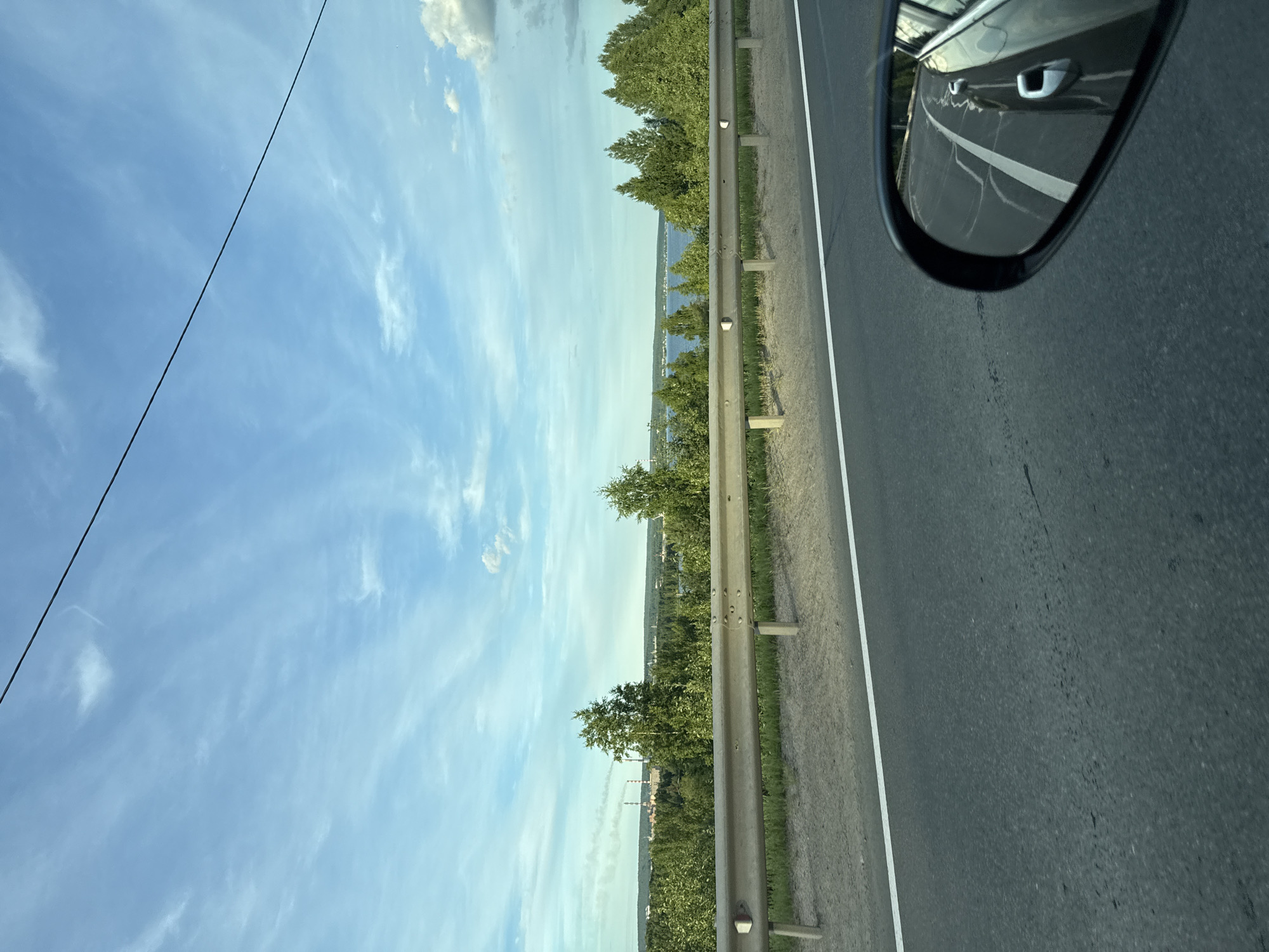 The image shows a view from a car traveling on a road, likely in or near Kondopoga, Russia, during daylight. The sky is blue with scattered white clouds and a black wire is visible stretching across the top of the frame. In the middle ground, a metal guardrail runs along the side of the road, separating it from a grassy embankment and a dense forest. Beyond the trees, a body of water is visible, possibly Lake Kondopoga or a river, and in the distance, industrial structures with smokestacks emitting smoke can be seen, indicating an industrial area within or near the city. A side-view mirror of the car is visible in the foreground on the right, reflecting the road and parts of the car.