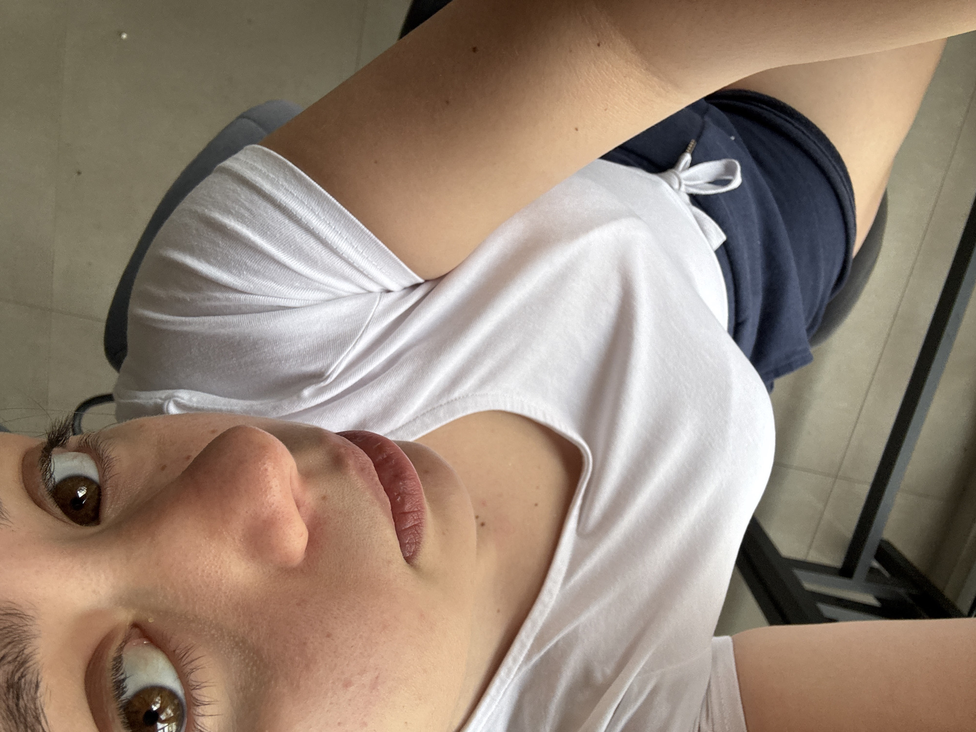 A selfie taken from a low angle, presumably by a person seated on a chair. The individual wears a white t-shirt and navy blue shorts with a white drawstring. Parts of a grey chair and black metal chair legs are visible. The environment is undefined and cannot be said to be in Garmen, Bulgaria.