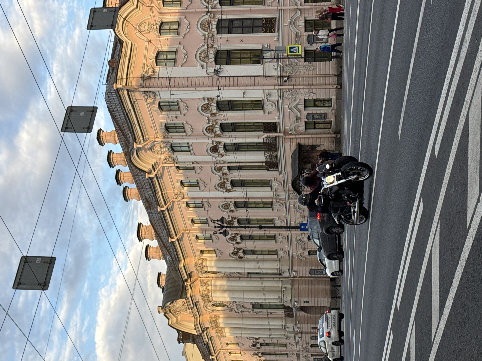 A view of a street in Saint Petersburg, Russia, featuring a large, ornate pink building with classical architectural elements. The building has numerous windows, decorative moldings, and prominent cornices. In the foreground, a motorcycle with two riders, both wearing helmets, is driving on the asphalt road. The road is marked with white lane lines and pedestrian crossing markings. Several cars are also present on the road, including a white sedan and a dark SUV. Pedestrians are visible on the sidewalk in front of the building. Overhead, a complex network of wires and two square signs suspended from them are visible against a cloudy sky. The lighting suggests it is late afternoon or early evening.
