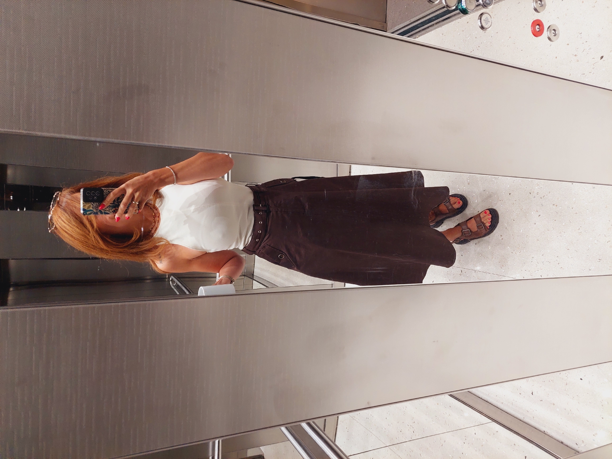 A woman with auburn hair is taking a mirror selfie in an elevator. She is wearing a white halter-neck top, a brown midi skirt with a belt, and brown sandals. She is holding a white mug in her left hand and her phone in her right hand. Her nails are painted red. The elevator walls are made of brushed metal, and the floor is speckled terrazzo. The elevator doors are partially open, revealing a glimpse of the outside, possibly a hallway or another part of the building. The woman is standing in what appears to be an elevator in a modern building, likely in the city of Sofia, Bulgaria.