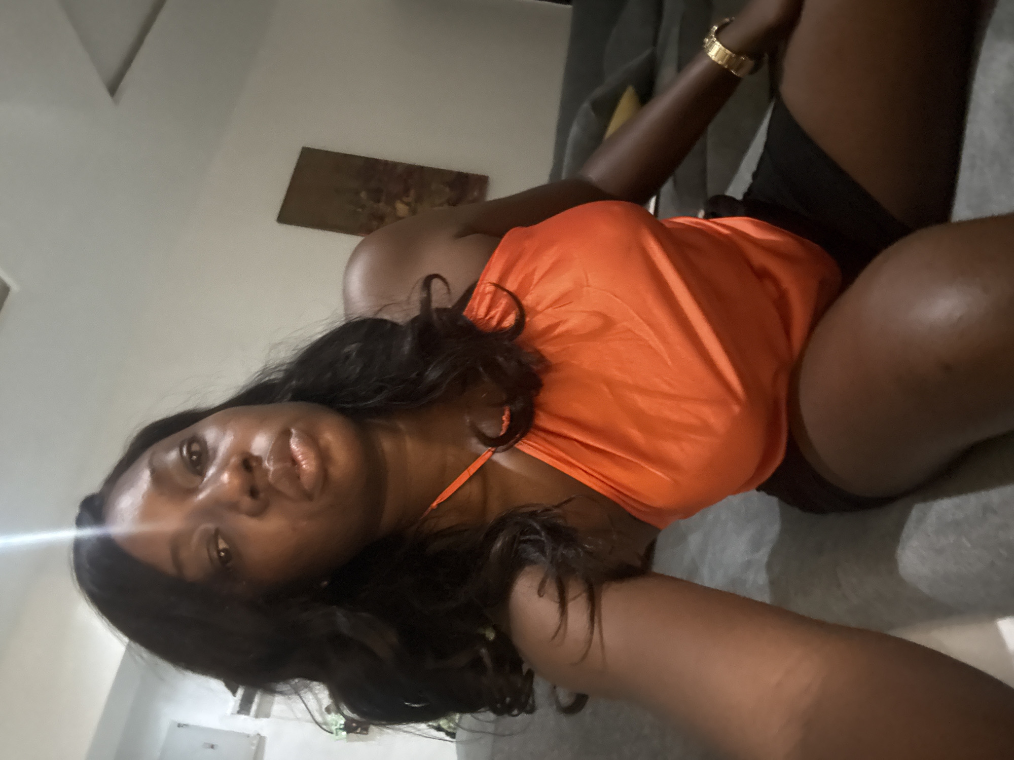 A dark-skinned woman with long, dark hair is lying on a gray surface. She is wearing an orange halter top and dark shorts. A gold watch is visible on her left wrist. A painting hangs on the wall in the background. The location is unspecified. The city of Iddo Tudunwada, Nigeria, cannot be confirmed as the location.