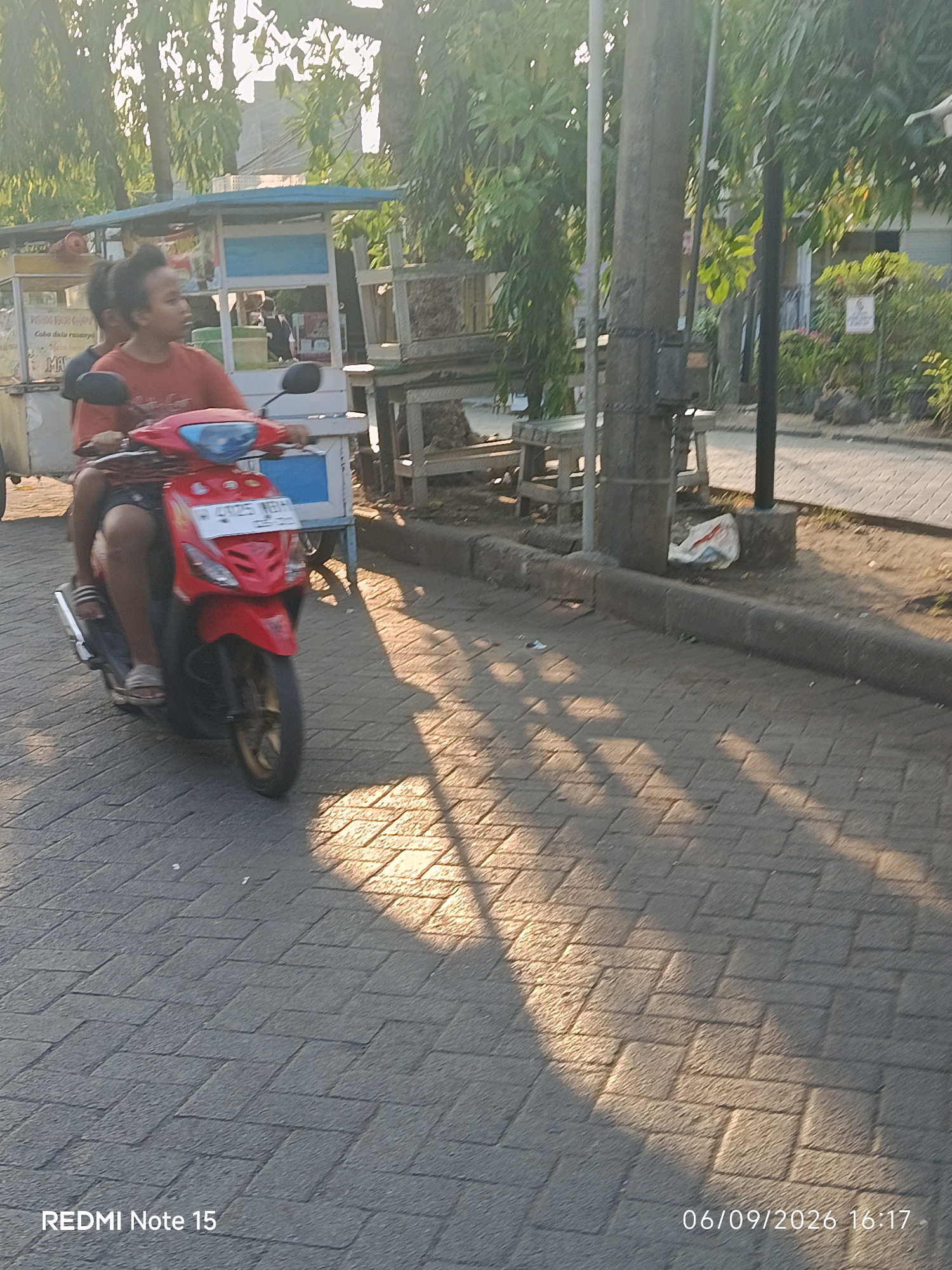 The image captures a scene in Keboansikep, Indonesia, with a clear emphasis on a red scooter in motion. Two individuals are visible on the scooter, with the person in front steering and the person behind appearing to be a child. They are riding on a paved walkway with a distinctive brick pattern. In the background, a food cart is present, suggesting a public area, possibly a street or market. Trees and foliage line the background, indicating an outdoor setting with natural elements. The lighting suggests it is either early morning or late afternoon, with the sun casting shadows on the walkway. The presence of the scooter and the people indicates activity and movement within the urban environment.