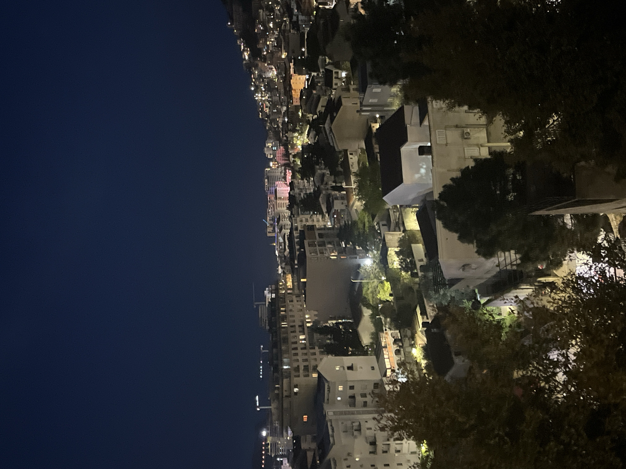 The image depicts a nighttime cityscape, likely of Budva, Montenegro, judging by the architecture and coastal location. The sky is a dark blue to black. Buildings of varying heights and architectural styles dominate the scene. Many buildings are illuminated with warm lights. There are trees visible along the right side of the frame. Overall, the image captures a scene of urban activity in a coastal city at night.