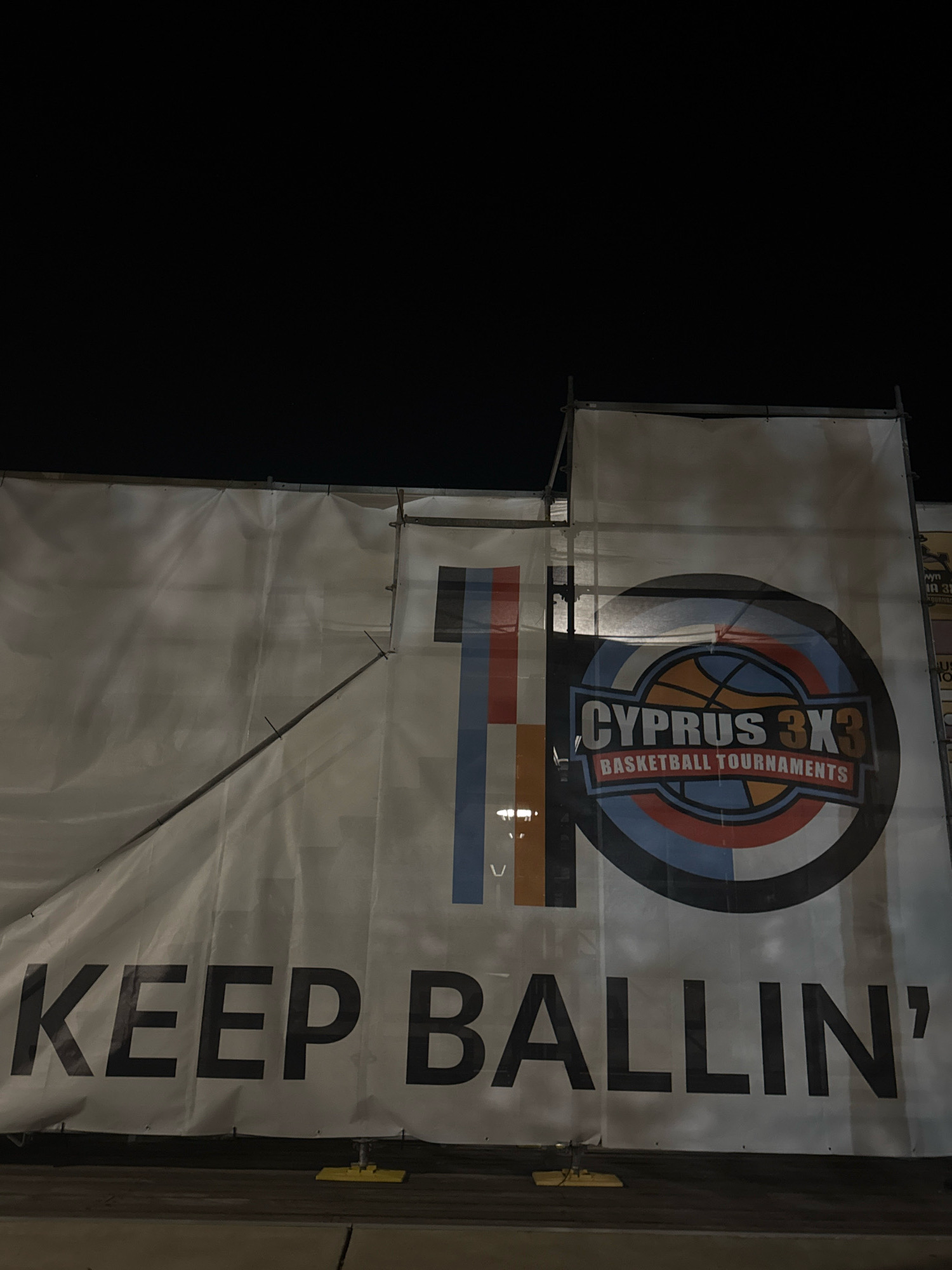 The image shows a large white banner displayed outdoors at night. The banner has a dark background with the text "KEEP BALLIN'" printed in large, bold, black capital letters across the bottom. Above this text, to the right, is a circular logo that reads "CYPRUS 3X3 BASKETBALL TOURNAMENTS" in smaller text. The logo features a stylized basketball in the center with various colored segments around it. To the left of the logo, there are vertical stripes of blue, orange, and white. The banner is supported by scaffolding. The lower portion of the image shows a dark ground, possibly asphalt or concrete, with yellow square anchors attached to the bottom of the banner. The overall scene suggests an outdoor event or advertisement in Limassol, Cyprus.