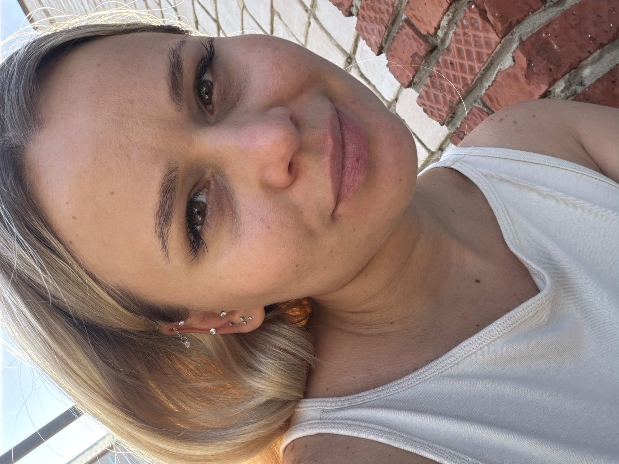 The image shows a woman with blonde hair, wearing a white tank top, with multiple ear piercings. She is positioned in front of a brick wall. It is not possible to determine the exact location in Sarapul, Russia, from the image alone.