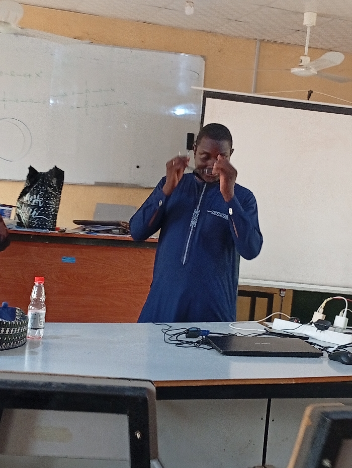 An African man in a navy blue tunic adjusts his glasses. He stands in a classroom with a whiteboard showing mathematical equations, and a projection screen. A table in front of him holds a laptop, a water bottle, and various wires and chargers. A ceiling fan is visible.