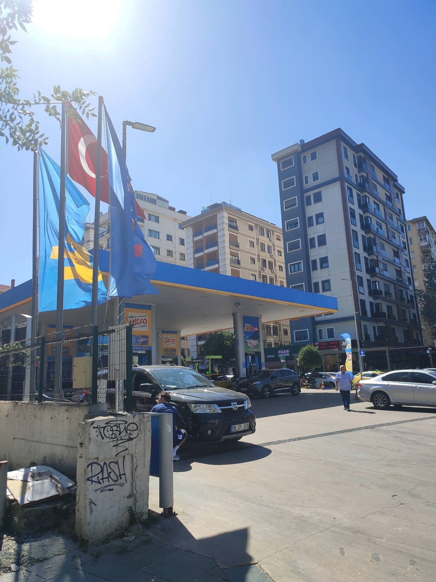 The image depicts a street scene in Kadıköy, Turkey, likely a commercial area. In the foreground, a concrete wall with graffiti is visible. Beyond this, a black SUV is parked under a gas station canopy, with a person in a blue and white striped shirt kneeling beside it. Flags, including a Turkish flag and blue flags, fly from poles near the canopy. Several other vehicles are present, including a silver sedan and a yellow car, as well as a green bus in the background. Apartment buildings of varying heights and architectural styles line the street, suggesting an urban environment. The sky is clear and blue, with strong sunlight casting shadows. The scene appears to be an active street with ongoing traffic and pedestrian activity.