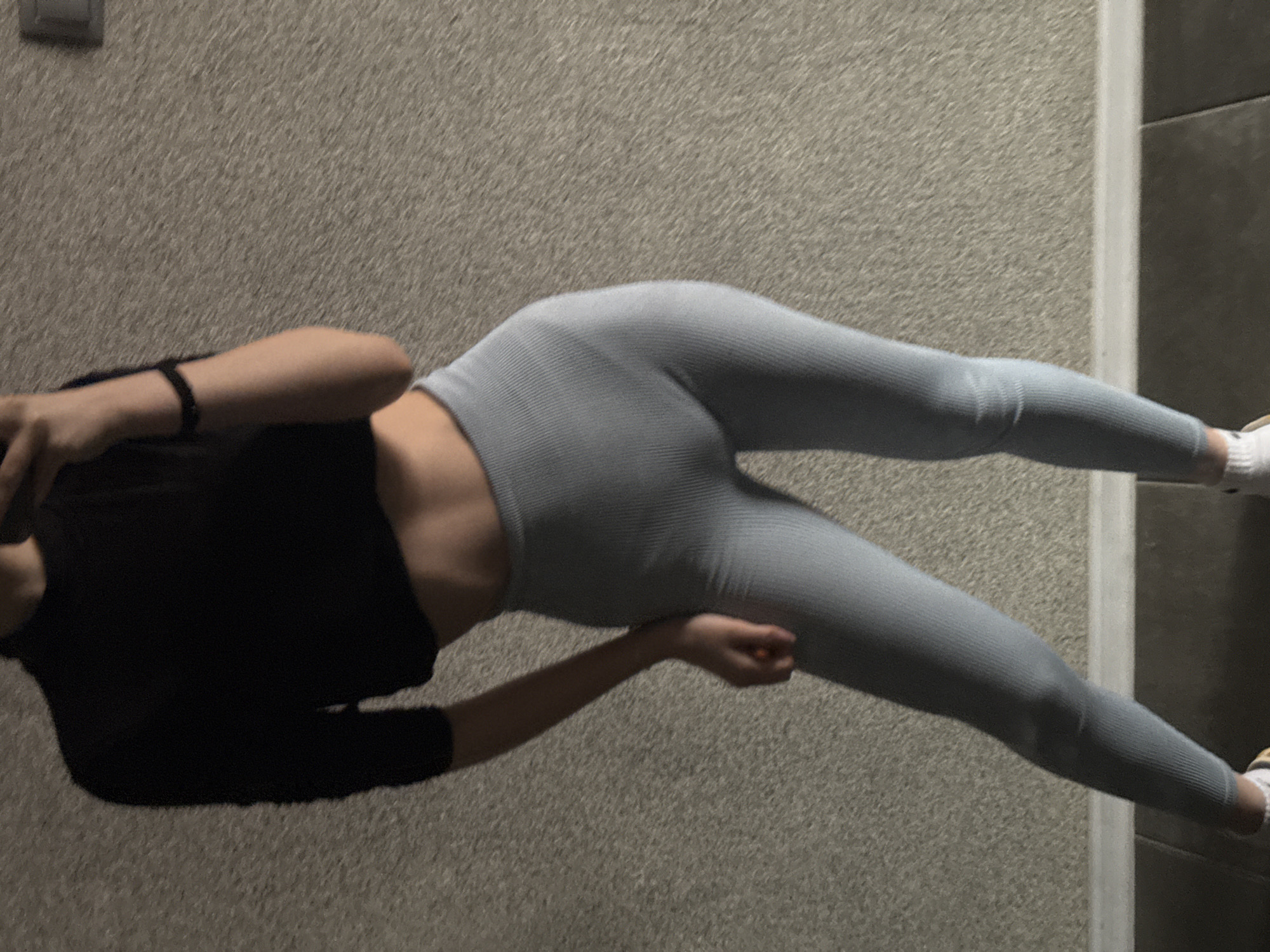A person is lying on a carpeted floor, wearing a black top and light gray leggings with white socks and athletic shoes. The person is oriented at an angle relative to the frame. The lighting suggests an indoor setting. A light switch plate is visible in the upper left corner. A white door frame is visible in the background to the right. The city of Sofia, Bulgaria, is not identifiable from this image.