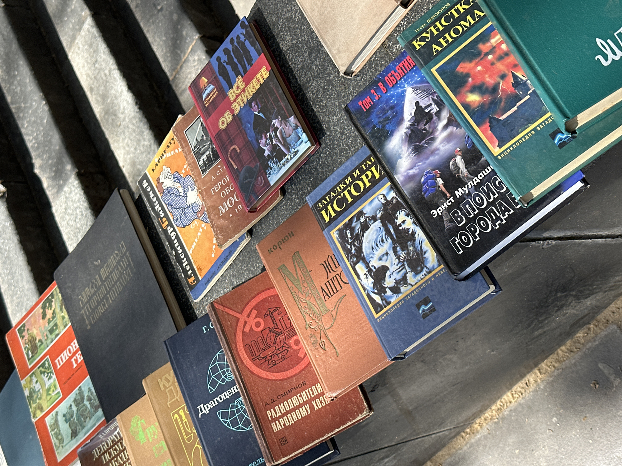 Numerous books, mostly with Cyrillic script, are displayed outdoors, likely on concrete or stone steps or a low wall. The strong shadows cast across the scene suggest bright daylight, possibly from the sun overhead, indicating an outdoor market or a street vendor's setup. The books are arranged somewhat haphazardly, piled and leaned against each other, suggesting casual browsing or sale.
The books vary in size, color, and apparent age. Several feature colorful covers with illustrations. Visible titles and authors include:
* A brown book with "РАДИОЛЮБИТЕЛИ НАРОДНОМУ ХОЗЯ" (Radio Amateurs for the National Economy) by А. Д. СМИРНОВ (A. D. Smirnov).
* An orange and brown book with "ГЕРОИ ОБО МОС 19." (Heroes of the Moscow Defense 19xx) and an illustration of a character.
* A red book titled "ВСЕ ОБ ЭТИКЕТЕ" (All About Etiquette) from a "ДОМАШНЯЯ КОЛЛЕКЦИЯ" (Home Collection).
* A blue book with "ЗАГАДКИ И ТА ИСТОРИ" (Mysteries and History).
* Another blue book titled "ТОМ З. В ОБЪЯТИЯ" (Volume 3. In Embrace/Grip).
* A prominent green book by Игорь ВИНОКУРОВ (Igor Vinokurov) titled "КУНСТКА АНОМА" (Kunstkamera Anoma), with a subtitle "ЭНЦИКЛОПЕДИЯ ЗАГАДОЧНОГО И НЕВЕ" (Encyclopedia of the Mysterious and Unkno...).
* A book by Эрнст Мулдаш (Ernst Muldashev) titled "В ПОИС ГОРОДА Е" (In Search of the City of...).
* Other books show partial text like "ПИОН ГЕ" (Pioneer Hero), "ДЕКОРАТ ИСКУС" (Decorative Art), and "Драгоцен" (Precious/Jewels).
Some books appear well-preserved, while others show signs of wear and tear, consistent with second-hand items. The setting implies a common street market scene, which can be found in cities like Tbilisi, Georgia.