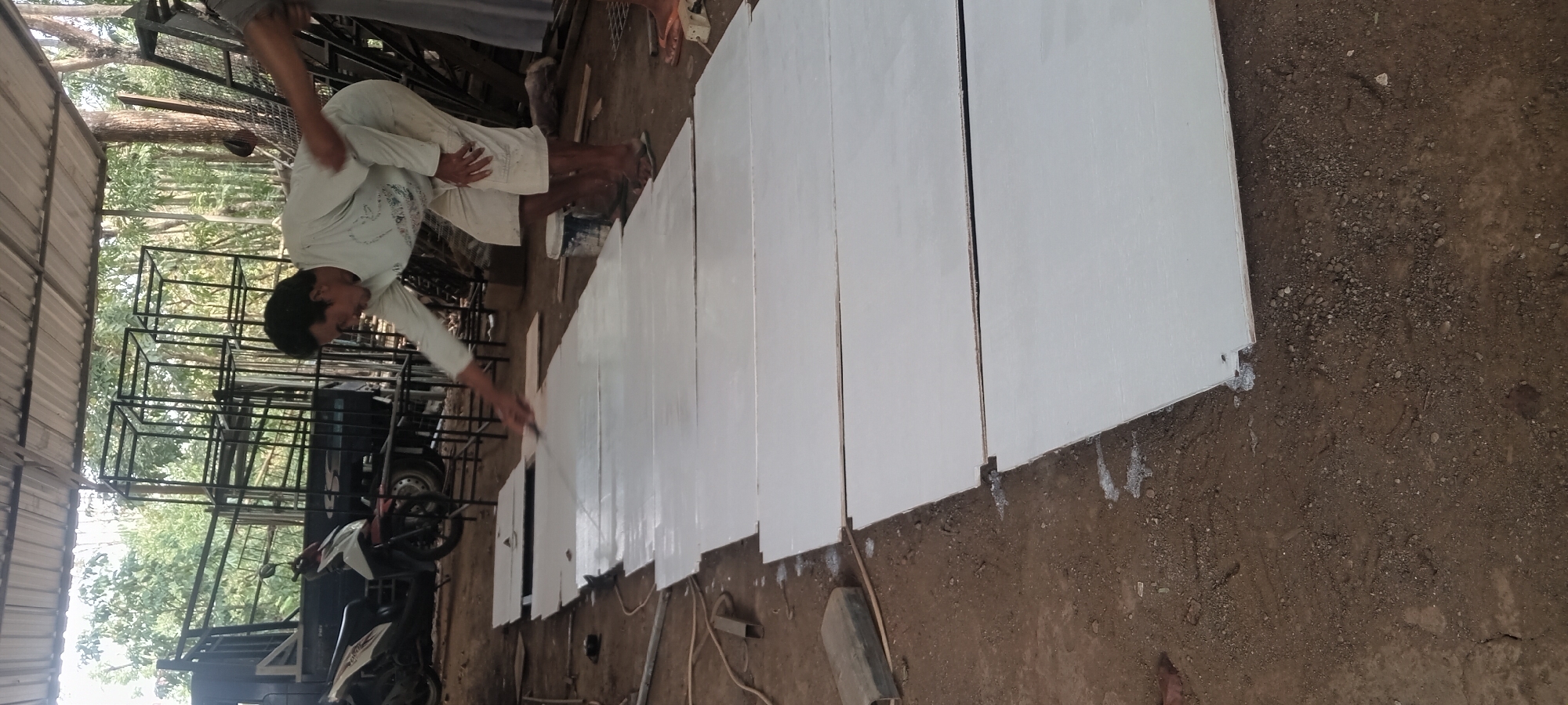 In Jogomulyo, Indonesia, within an open-air workshop covered by a corrugated metal roof, a man is painting several rectangular white panels laid out on the dirt floor. He is wearing a light-colored long-sleeved shirt and shorts, and he is using a brush. To the left of the panels, a motorcycle is parked next to a black truck with a metal frame structure on its bed. In the background, more metal structures and trees are visible, suggesting an outdoor industrial or construction setting. A can of paint is placed near the man's feet.
