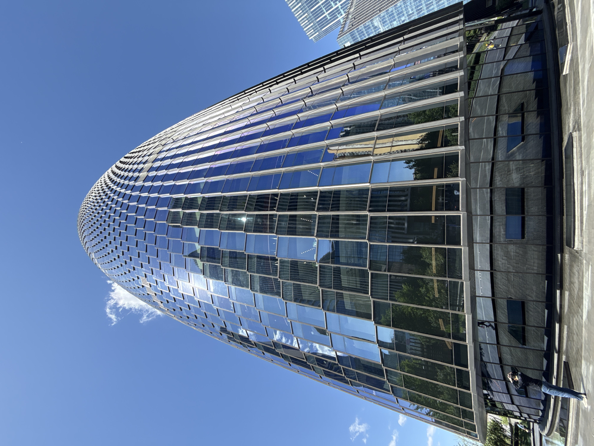 A low-angle shot captures a curved glass building against a clear blue sky with a few wispy clouds. The building's facade consists of numerous small, reflective square panels, creating a textured, mosaic-like appearance. The panels reflect the sky, other buildings, and trees, adding depth and complexity to the surface. On the right side of the building, a dark, rectangular section contrasts with the reflective panels. In the lower right corner, a person wearing jeans is partially visible, holding a camera pointed towards the building. Another taller building is seen in the background. The photo appears to be taken in Fengtai District, China.