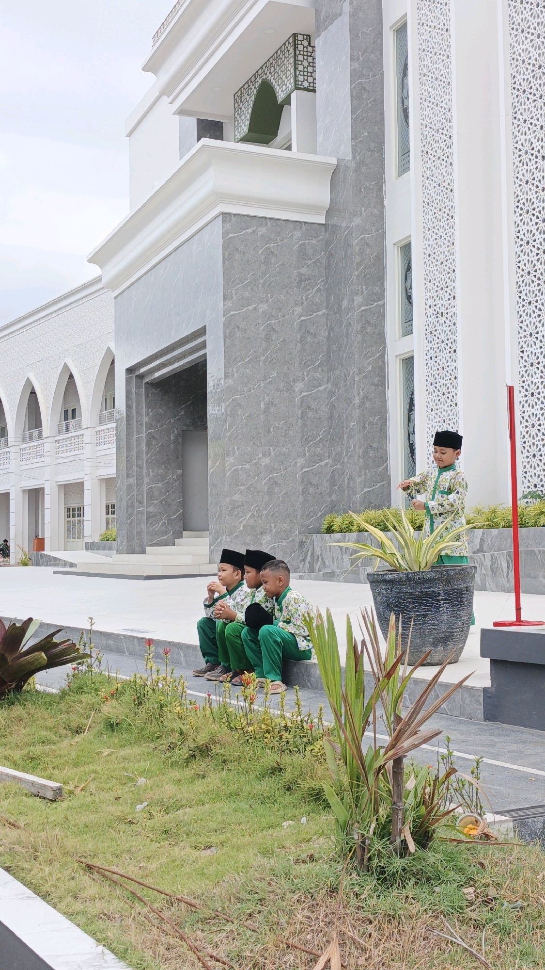 The image shows four young boys dressed in traditional Islamic attire, sitting and standing in front of a modern mosque. Three boys are sitting on a gray stone ledge, while one boy stands slightly to the right. They are all wearing black kufis (caps) and green shirts with white patterns. The mosque features a white facade with intricate geometric designs and gray marble accents. A large potted plant with spiky leaves is placed near the standing boy. The scene appears to be in a public outdoor area, with some grass and landscaping visible in the foreground. The sky is overcast. The image appears to be taken in the city of Desa Karangbenda, Indonesia, given the architectural style and attire.