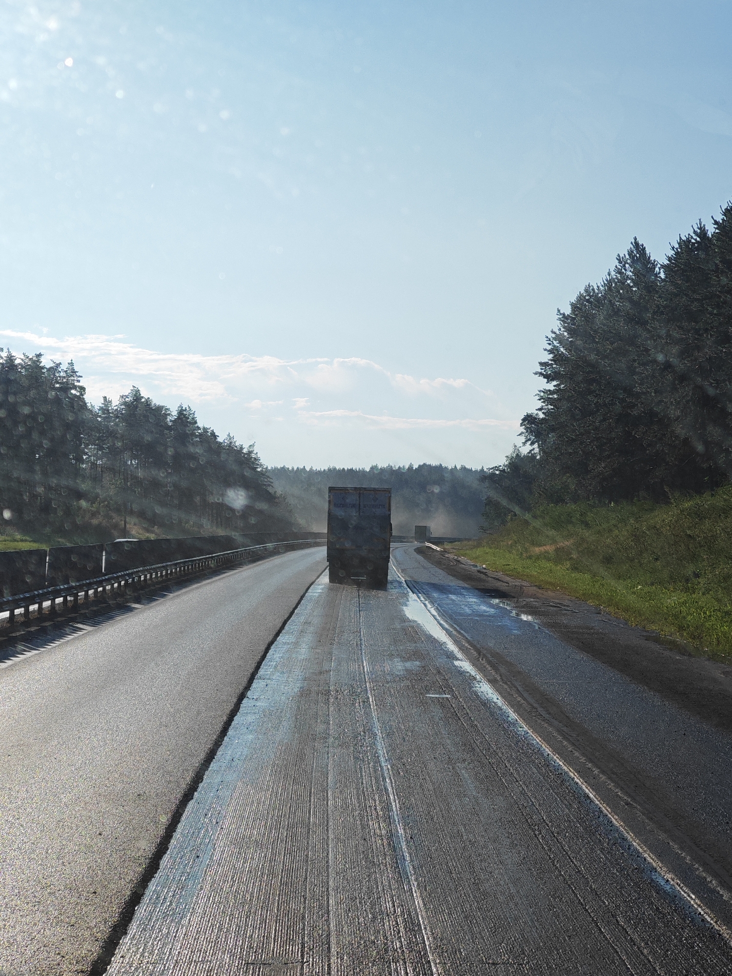 The image captures a highway scene on a sunny day, with a clear blue sky dotted with light clouds. The road appears to be undergoing resurfacing. The foreground shows a freshly paved lane with a textured surface, likely due to recent work. Further down the road, a truck is visible, emitting a faint trail of smoke or dust. On the right side of the road, a dense line of coniferous trees forms a natural barrier, suggesting a rural or semi-rural location. A guardrail runs along the left side of the highway, separating the lanes and the embankment beyond. The overall impression is one of ongoing infrastructure work on a highway surrounded by nature. This scene is located on the highway near Kurumoch, Russia.