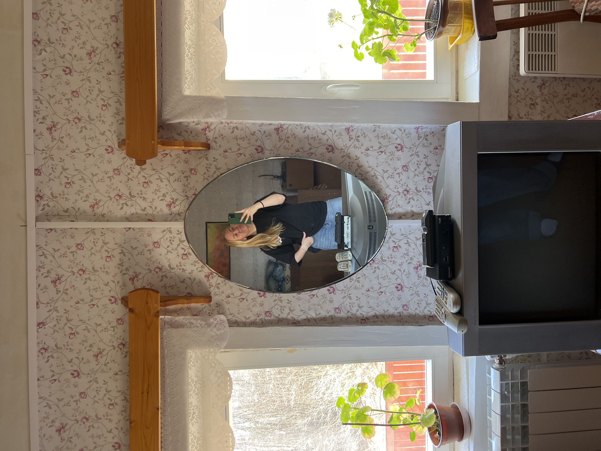 A woman stands in an indoor location, reflected in an oval mirror. She holds a phone, presumably taking the picture. The mirror is mounted on a wall covered with patterned wallpaper. Beside the mirror is a television and several remote controls. Natural light enters through a window near a potted plant. The setting has floral wallpaper. Based on visual analysis and the presence of Russian text, the image may be taken at Чернышово, Russia.