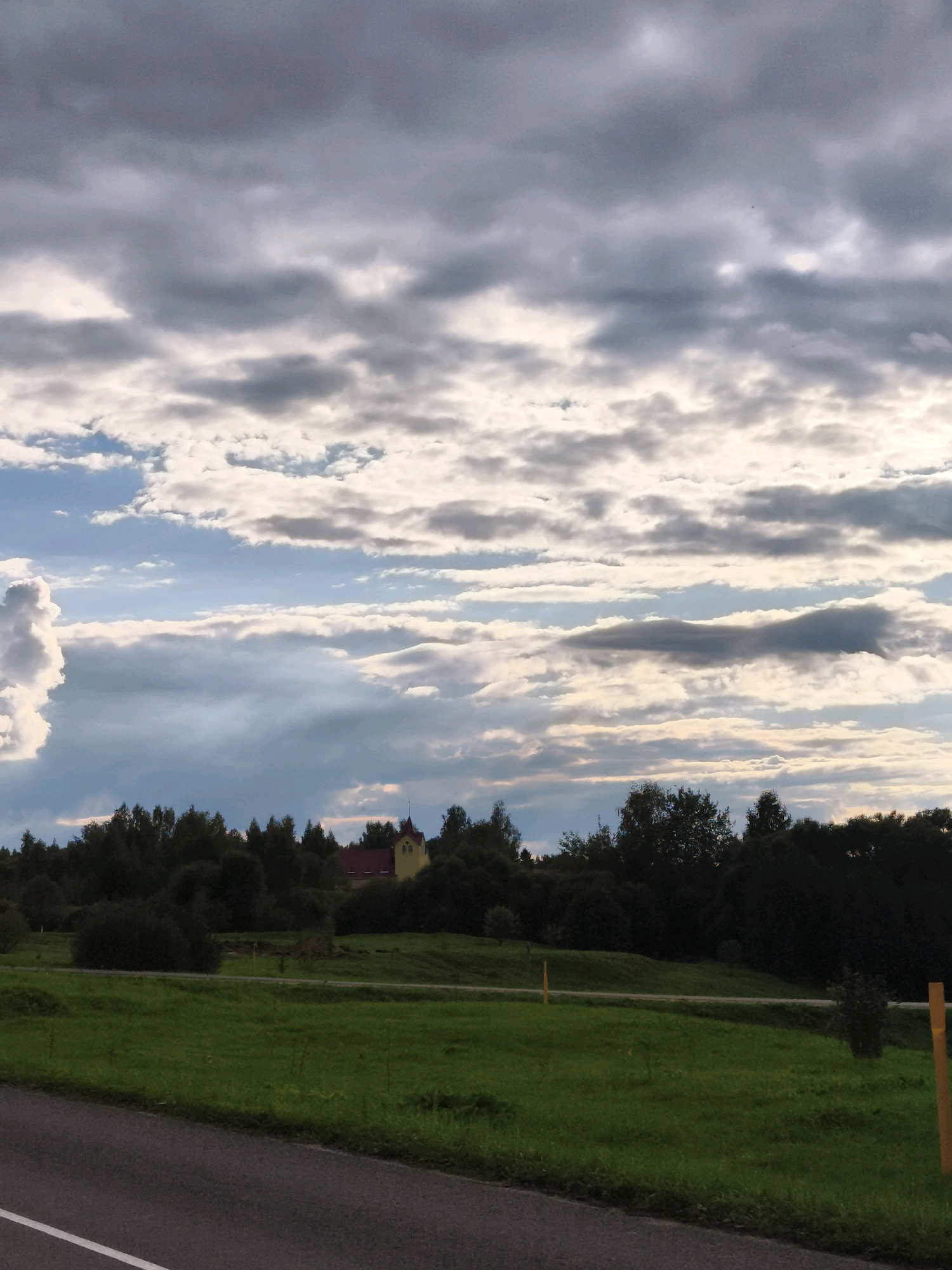 The image shows a rural landscape in the vicinity of Górski Selski Sovet, Belarus. The foreground features a paved road with a white line marking its edge, angling towards the bottom left. Beyond the road, a lush green field stretches into the middle ground, dotted with sparse vegetation. Further back, a dense line of dark green trees forms a natural barrier across the horizon. Nestled within this tree line, a building with a yellow facade and a red roof is visible. The sky above is a dramatic display of clouds, with patches of blue peeking through. Heavy, grey clouds dominate the upper portion of the frame, creating a sense of overcast weather, while lighter, billowy clouds appear lower in the sky, some illuminated by sunlight. Two yellow posts are visible in the field, one in the middle ground and another on the right edge of the frame, likely serving as markers or property boundaries. The overall scene depicts a tranquil, natural environment under a dynamic sky.