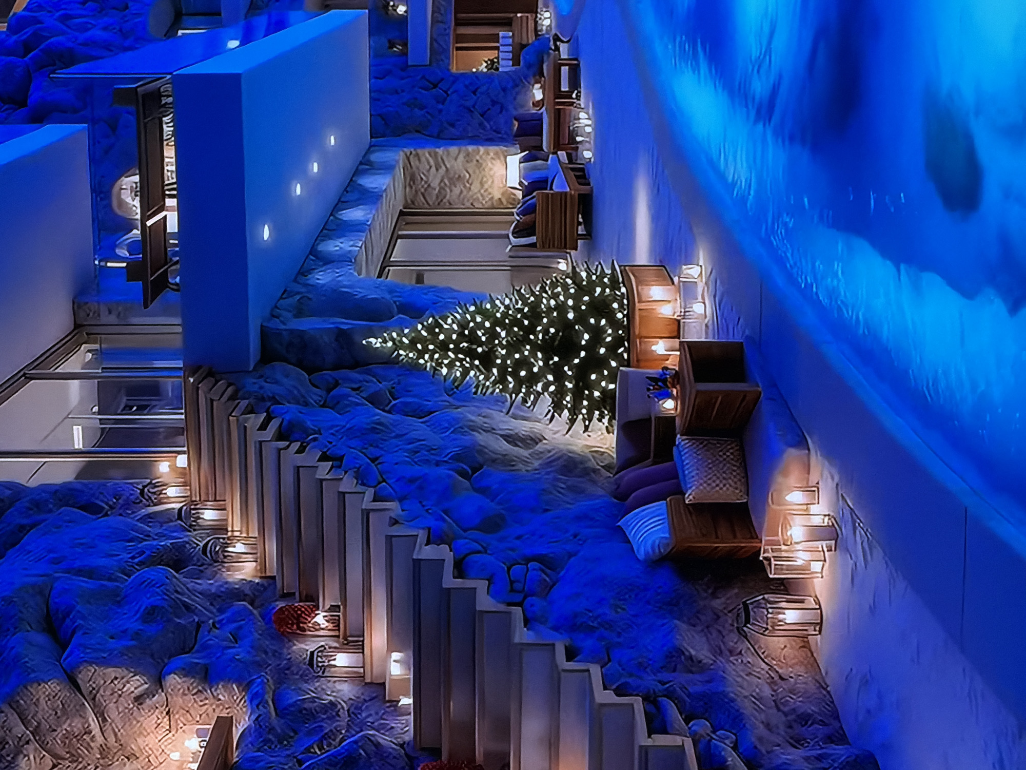 This image captures a uniquely designed interior space, likely a hotel lobby, lounge, or relaxation area, located in Sofiivska Borshchahivka, Ukraine. The scene is dominated by a pervasive, cool blue lighting that bathes most surfaces, creating an ethereal and tranquil ambiance.
The setting features organic, sculpted architecture with undulating, rock-like textures that form the ground and lower walls. A prominent feature is a brightly lit Christmas tree, adorned with numerous white lights, positioned on a lower level, indicating a festive holiday season.
Various recessed seating areas are integrated into the textured landscape, furnished with cushions in shades of blue and white stripes. Some of these seating alcoves include wooden shelving units that hold clear, lantern-style light fixtures emitting a warm, inviting glow, contrasting with the dominant blue light.
Along the left, a long, rectilinear white structure, possibly a balustrade or architectural element, is punctuated by several small, circular white lights. On the right, a tall wall is intensely illuminated with blue light, creating a dramatic, almost icy or watery effect. Pathways and steps, partially bordered by wooden railings, are visible, navigating the contoured terrain. Glass partitions or barriers are also part of the modern design. The scene is devoid of people, contributing to the serene and composed atmosphere. The artificial lighting suggests the scene takes place during the evening or night. There is no visible text.
